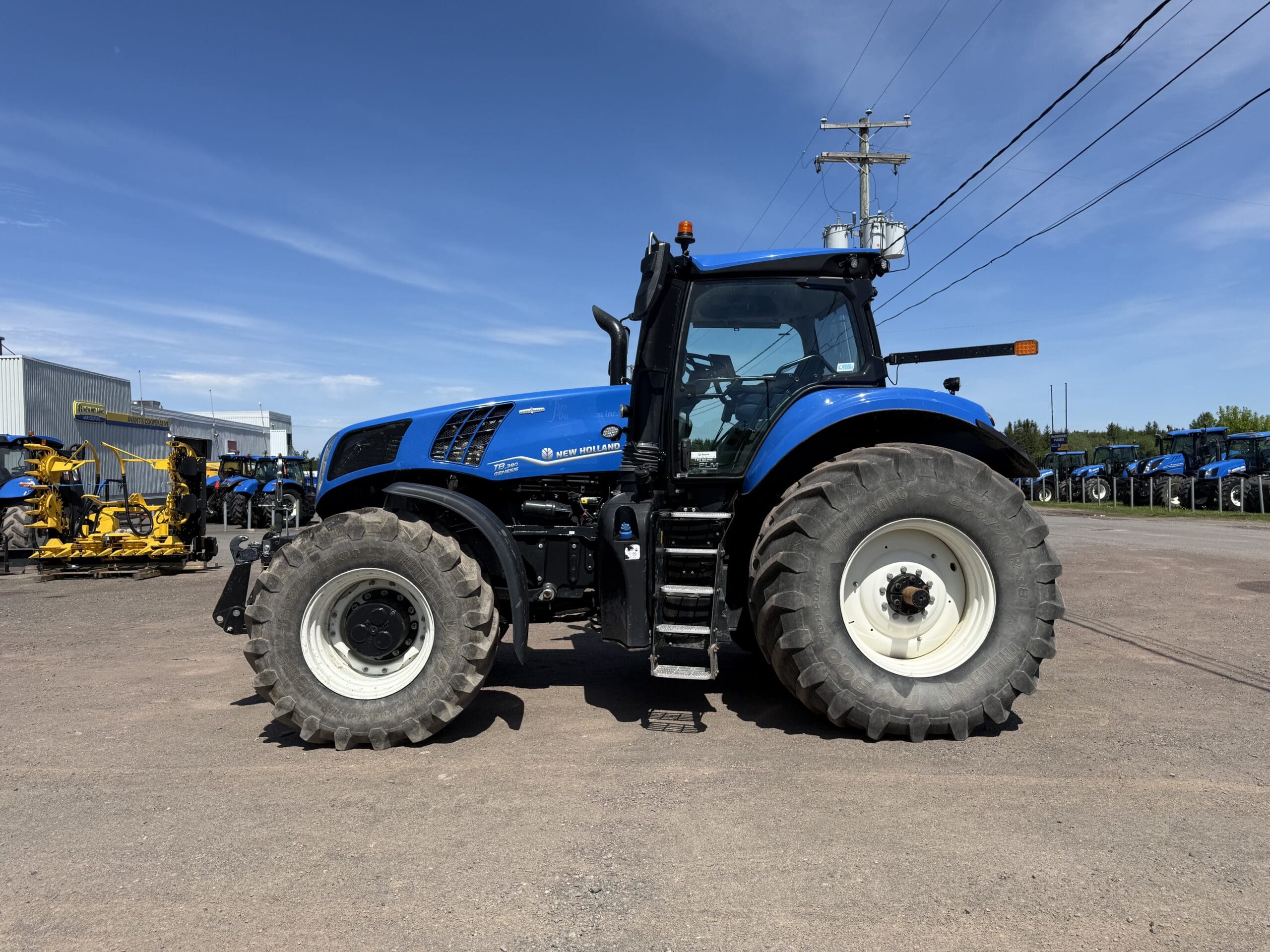 Un grand tracteur bleu aux roues noires et blanches est garé sur un terrain en gravier, sous un ciel d'un bleu limpide. D'autres machines et équipements sont visibles à l'arrière-plan.