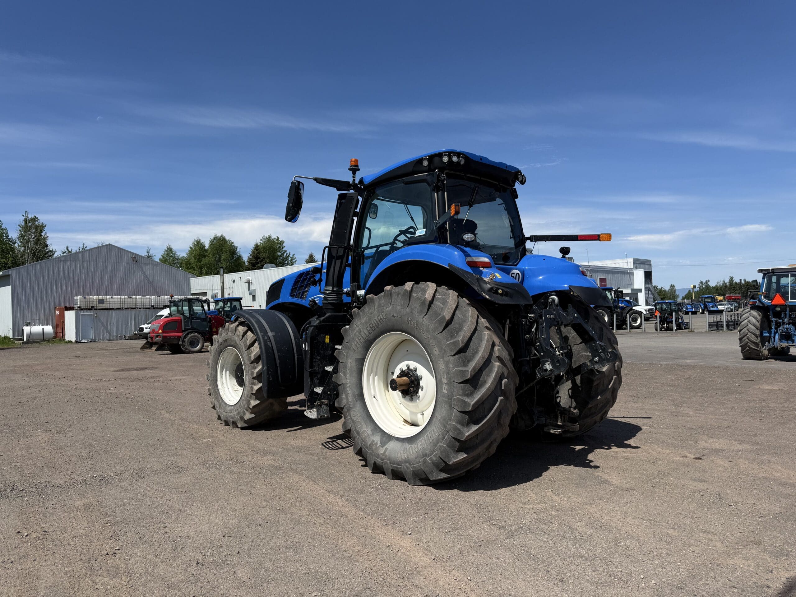 Un grand tracteur bleu équipé de pneus surdimensionnés est garé sur un terrain en terre battue sous un ciel d'un bleu limpide ; on aperçoit en arrière-plan des bâtiments, des arbres et d'autres véhicules.