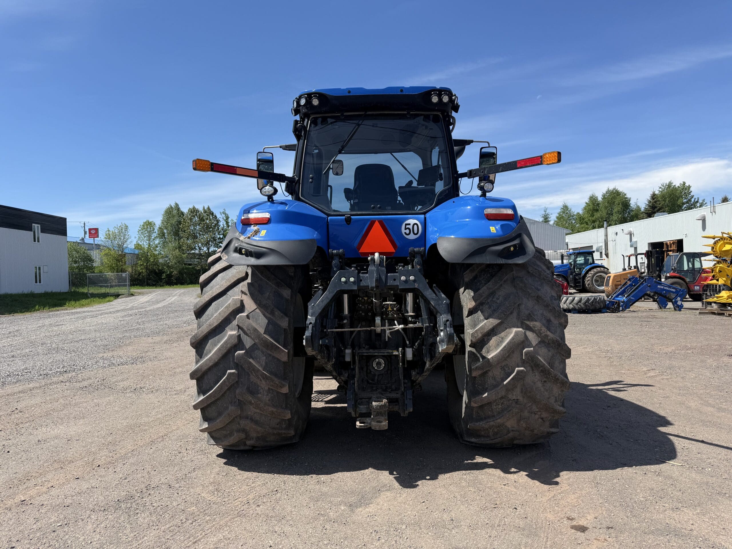 Un tracteur bleu est garé sur un terrain en terre battue, vu de l'arrière. On distingue ses gros pneus arrière boueux, ses catadioptres orange et son panneau d'avertissement triangulaire. Des bâtiments et d'autres engins apparaissent à l'arrière-plan sous un ciel dégagé.