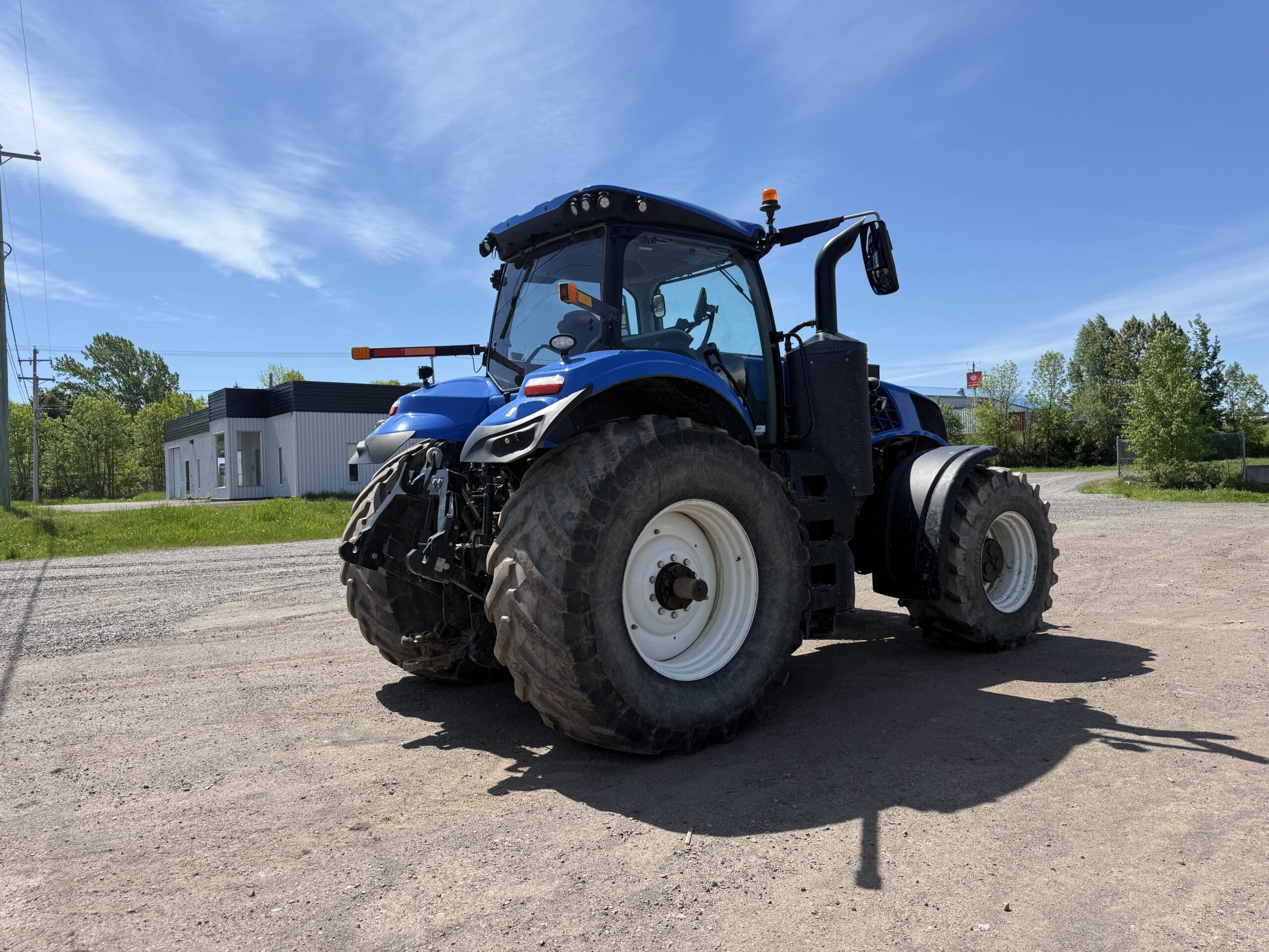 Un tracteur bleu équipé de gros pneus est garé sur un terrain en terre battue, sous un ciel bleu limpide, avec quelques bâtiments et des arbres verts visibles à l'arrière-plan.