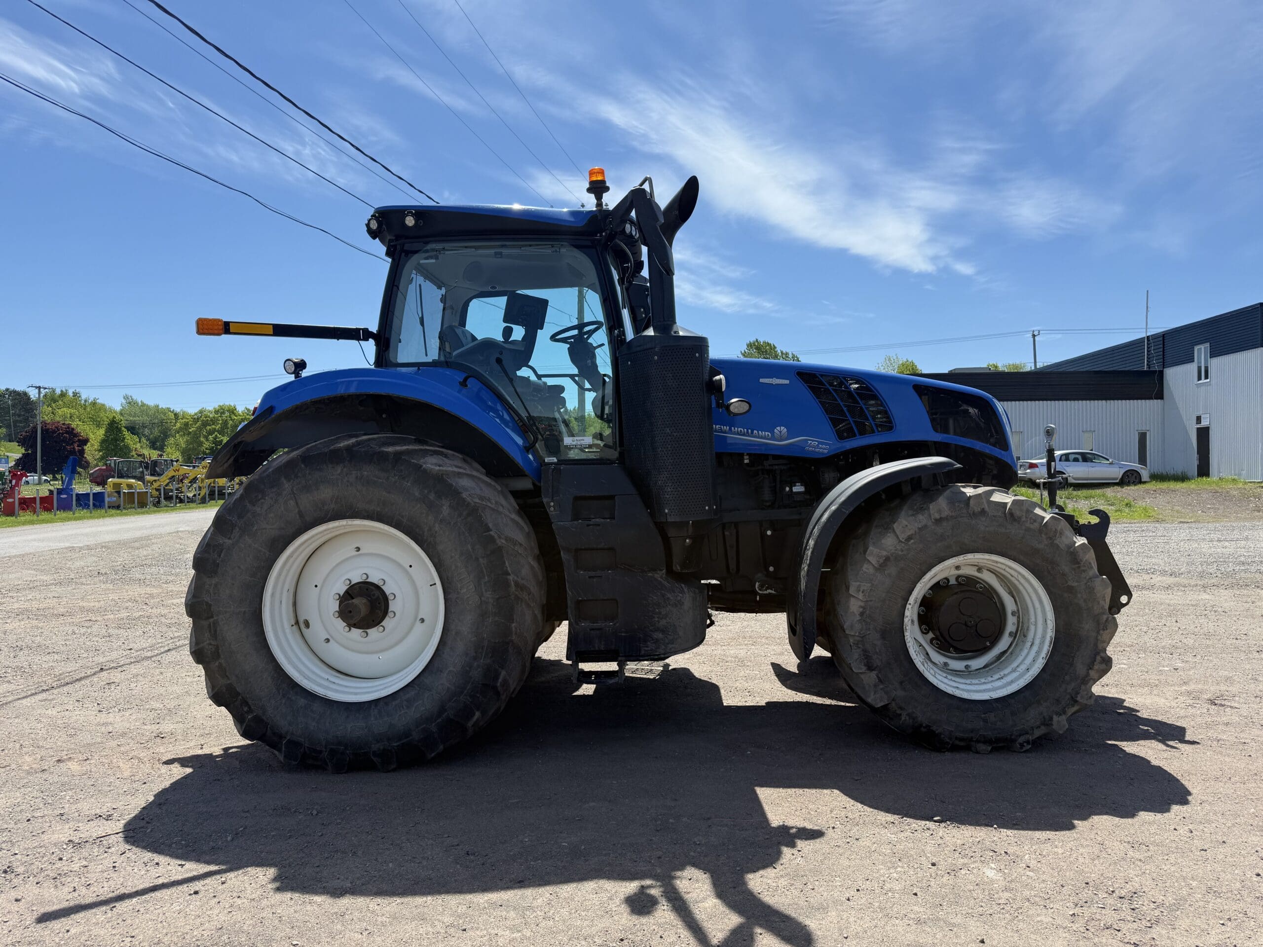 Un tracteur bleu équipé de gros pneus est garé sur un terrain en gravier, sous un ciel d'un bleu éclatant, avec un bâtiment blanc et des arbres verts en arrière-plan.