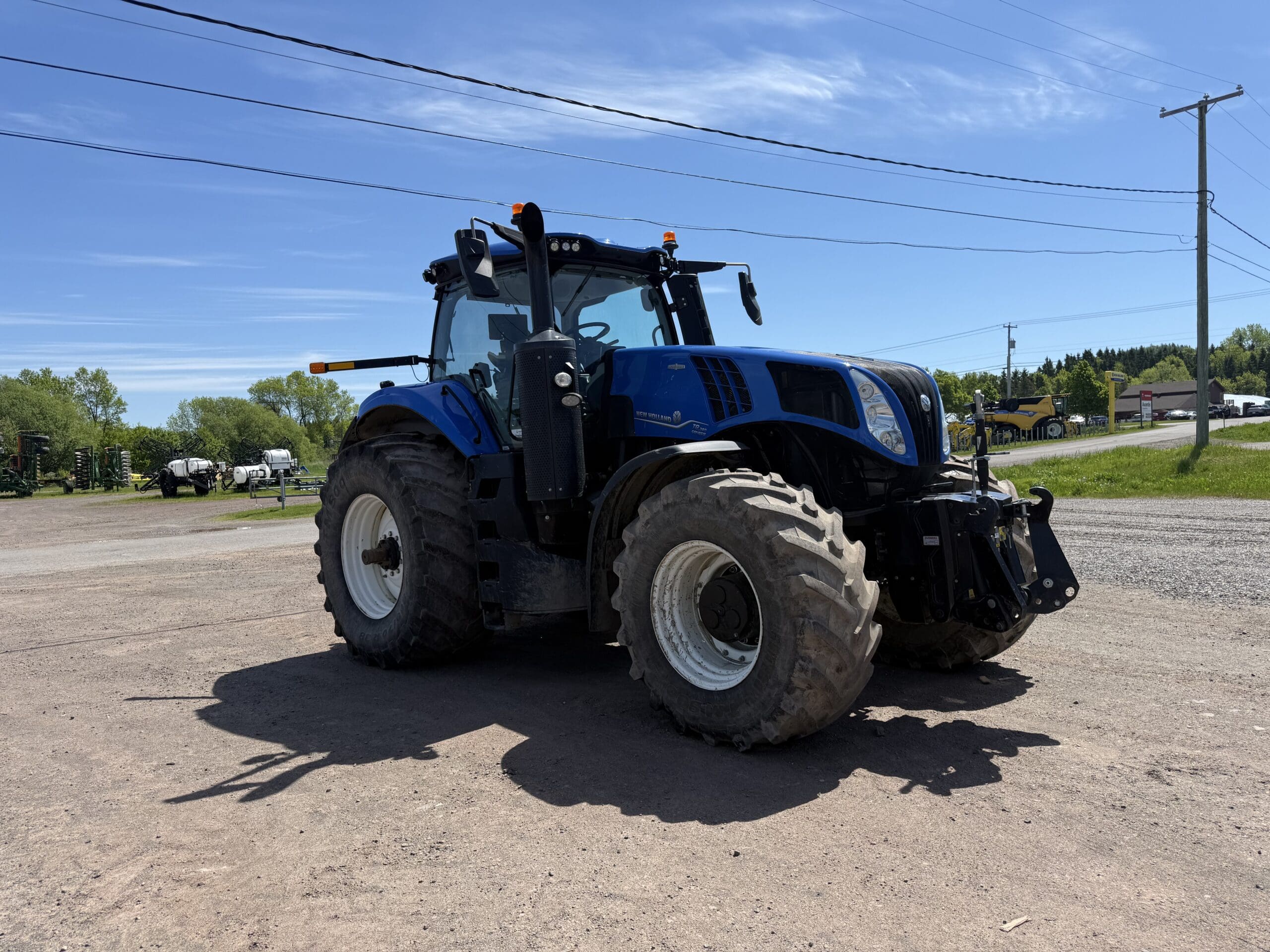 Un gros tracteur bleu équipé de pneus surdimensionnés est garé sur un terrain en gravier, sous un ciel clair et ensoleillé. On aperçoit des arbres et des poteaux électriques à l'arrière-plan.