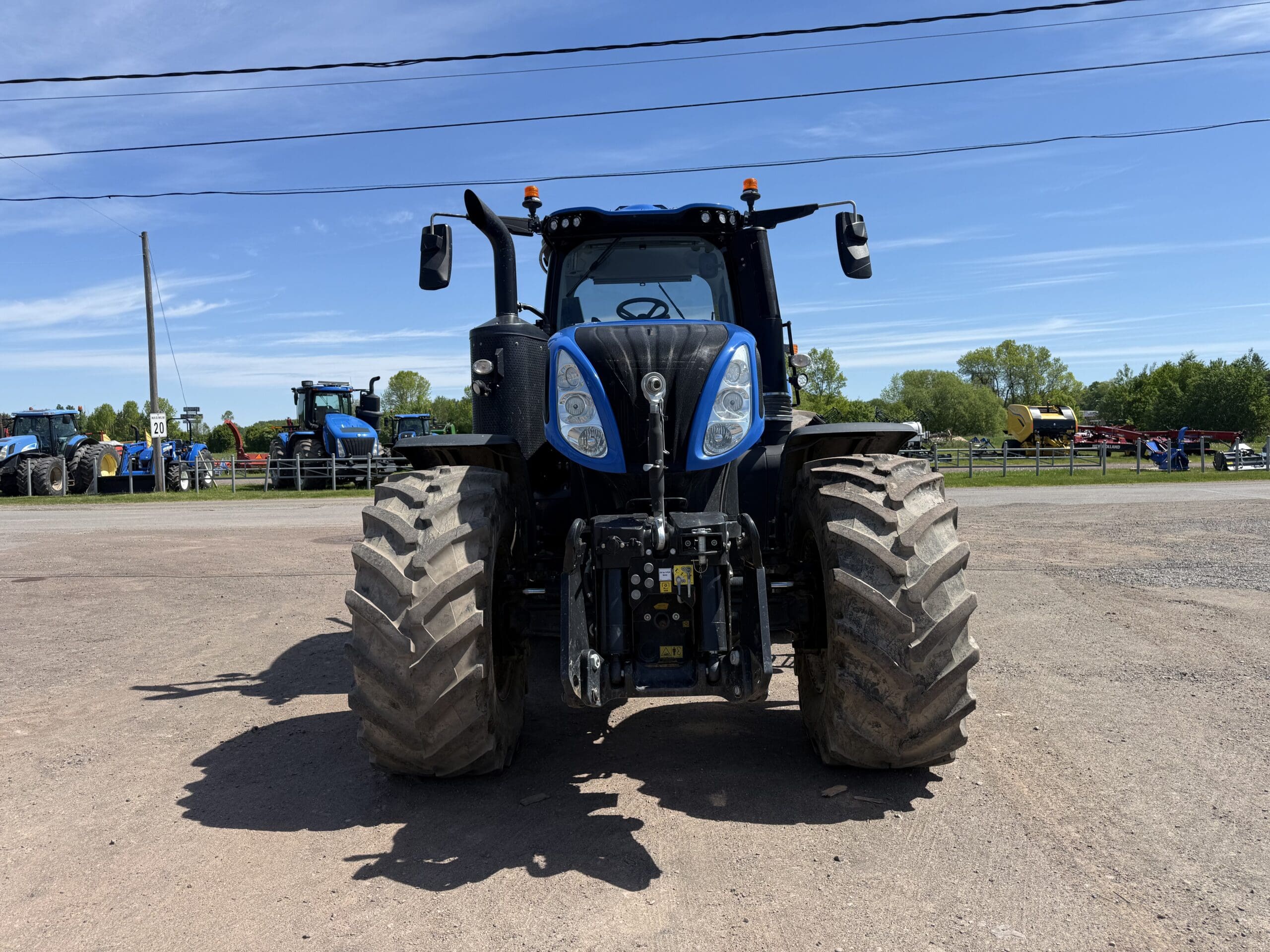 Un grand tracteur bleu équipé de larges pneus avant est garé sur un terrain en terre battue sous un ciel d'un bleu limpide, avec du matériel agricole et de la verdure en arrière-plan.