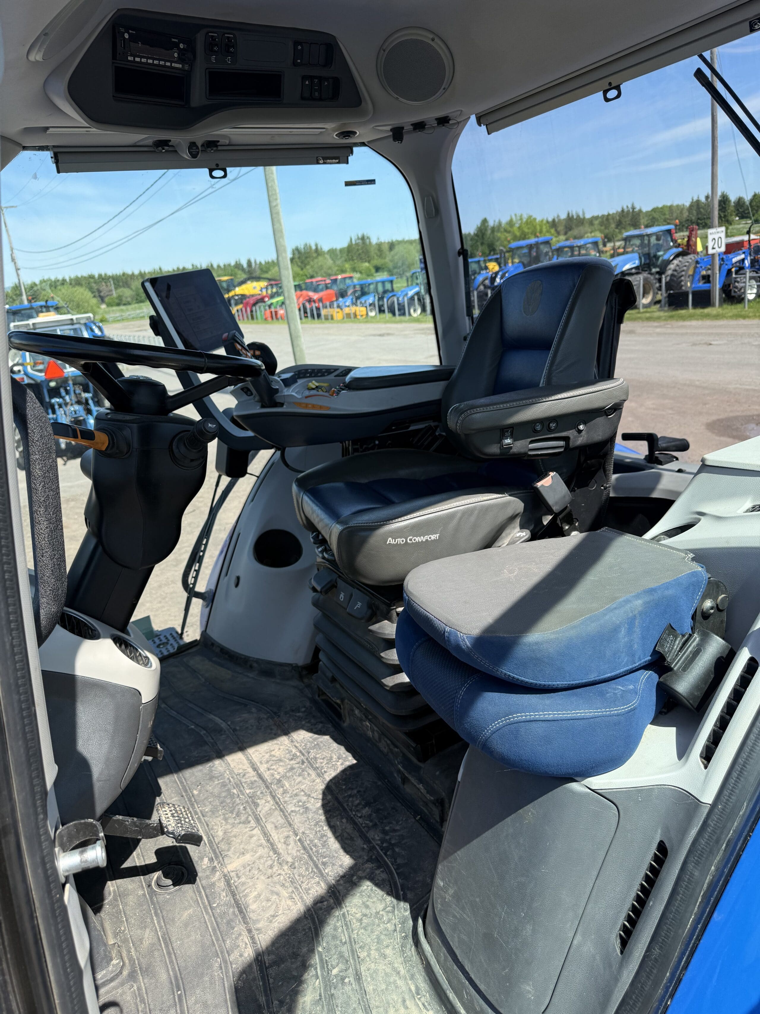 Intérieur de la cabine d'un tracteur montrant le siège du conducteur, les commandes, le volant et un siège passager bleu. La cabine est dotée de grandes vitres, à travers lesquelles on aperçoit d'autres tracteurs et le paysage extérieur.