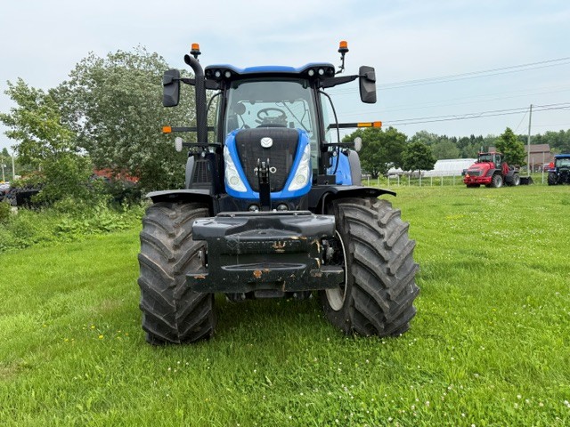 Un tracteur bleu est garé sur l'herbe verte, face à l'avant. À l'arrière-plan, sous un ciel nuageux, on aperçoit des arbres, un autre tracteur et une serre au loin.