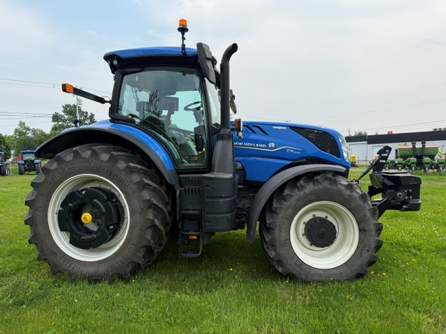 Un tracteur bleu équipé de gros pneus noirs est garé sur l'herbe verte. Le ciel est nuageux et on aperçoit du matériel agricole à l'arrière-plan.
