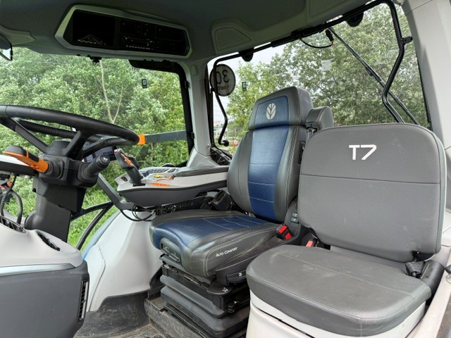 Intérieur de la cabine d'un tracteur, où l'on distingue deux sièges, dont l'un porte l'inscription « T7 », un volant, un tableau de bord équipé de leviers et de boutons, ainsi que de grandes vitres donnant sur des arbres verdoyants à l'extérieur.