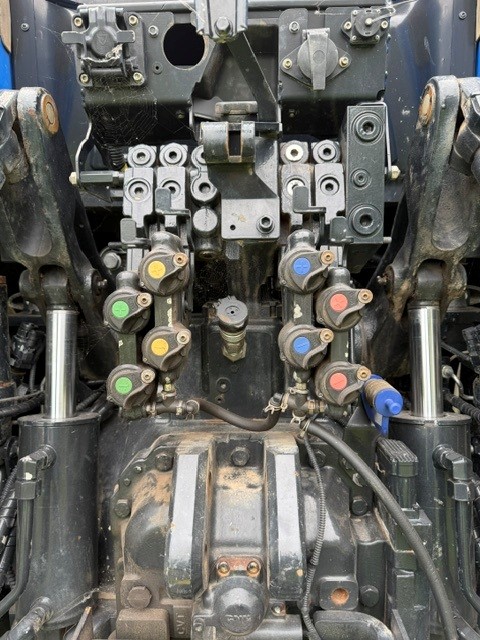 Gros plan sur le tableau de raccordement hydraulique et électrique d'un tracteur, comportant plusieurs capuchons identifiés par des codes couleur — vert, jaune, rouge et bleu —, entourés de pièces métalliques, de leviers et de flexibles hydrauliques.