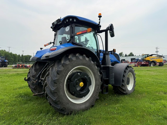 Un grand tracteur bleu est garé sur une pelouse verte ; on le voit de dos, du côté gauche. Le ciel est nuageux et on aperçoit du matériel agricole à l'arrière-plan.