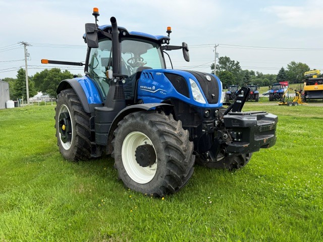 Un grand tracteur bleu aux roues blanches est garé sur une pelouse verte, avec des arbres et des poteaux électriques en arrière-plan, sous un ciel partiellement nuageux.