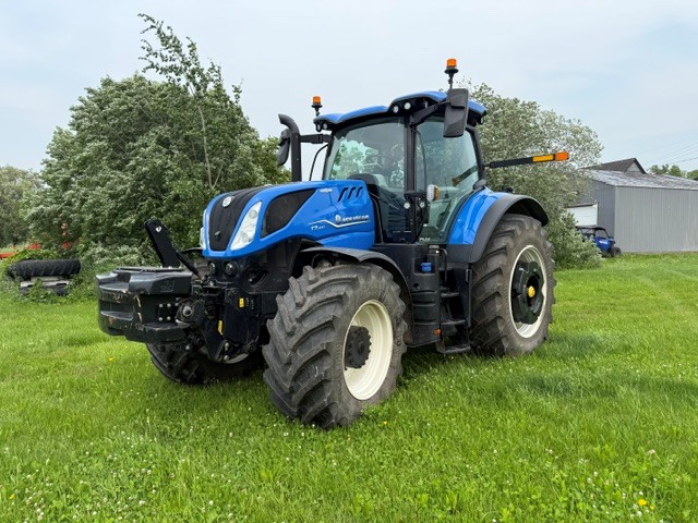 Un grand tracteur bleu aux roues noires et blanches est garé sur l'herbe verte, près d'arbres et d'un bâtiment gris, par une journée nuageuse.