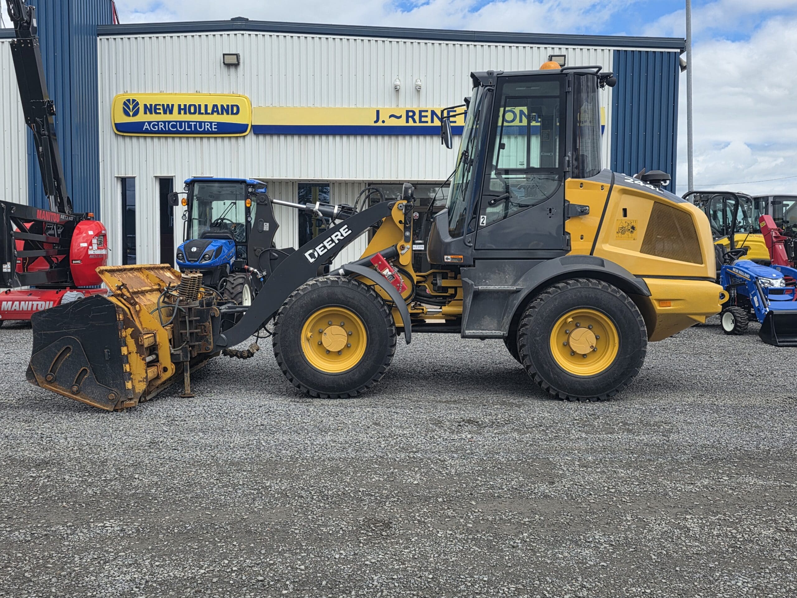 Une chargeuse frontale John Deere jaune équipée d'un godet est garée sur du gravier devant un bâtiment de New Holland Agriculture, par une journée partiellement nuageuse.