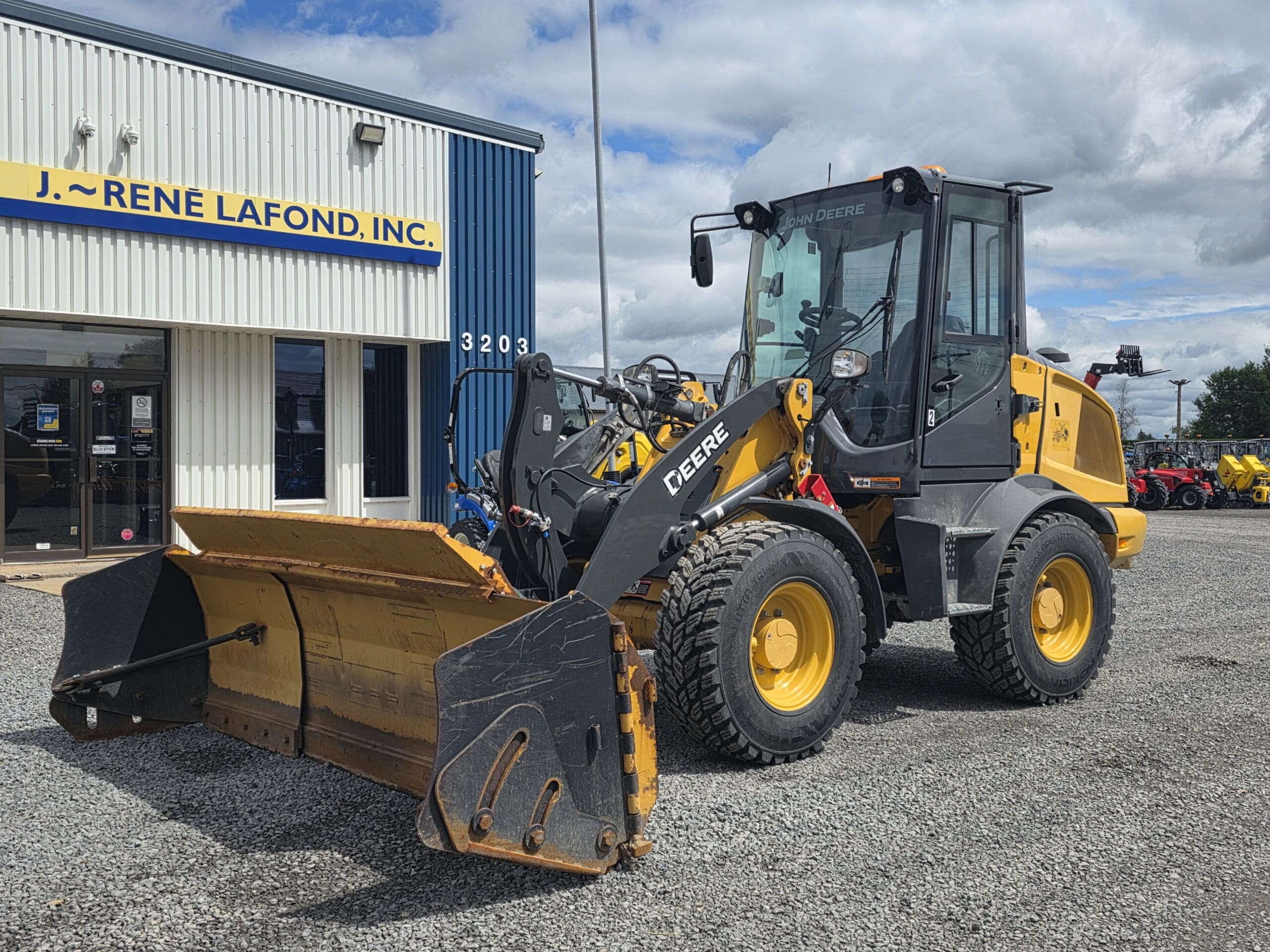 Une chargeuse frontale John Deere jaune est garée sur du gravier devant un bâtiment industriel bleu et blanc sur lequel on peut lire l'enseigne « J. René Lafond, Inc. ».