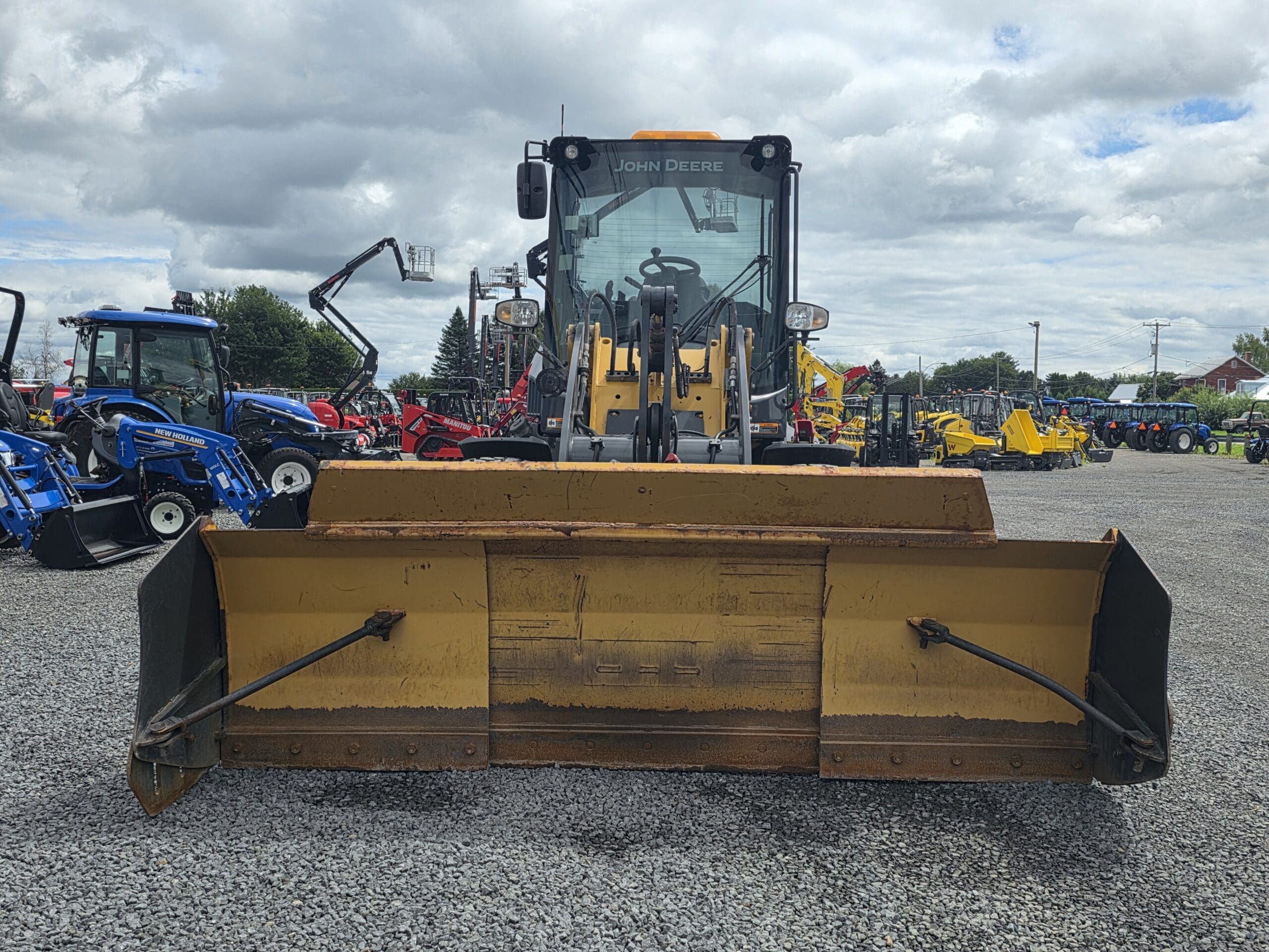 Un chargeur frontal John Deere jaune équipé d'une grande lame est garé sur du gravier, entouré de divers autres tracteurs et engins, par une journée nuageuse.