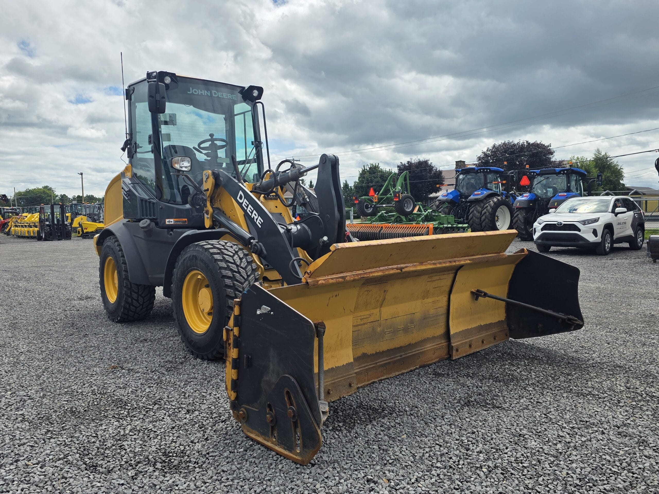 Une chargeuse frontale John Deere jaune, équipée d'une grande lame, est garée sur du gravier, entourée d'autres machines et véhicules, sous un ciel nuageux.