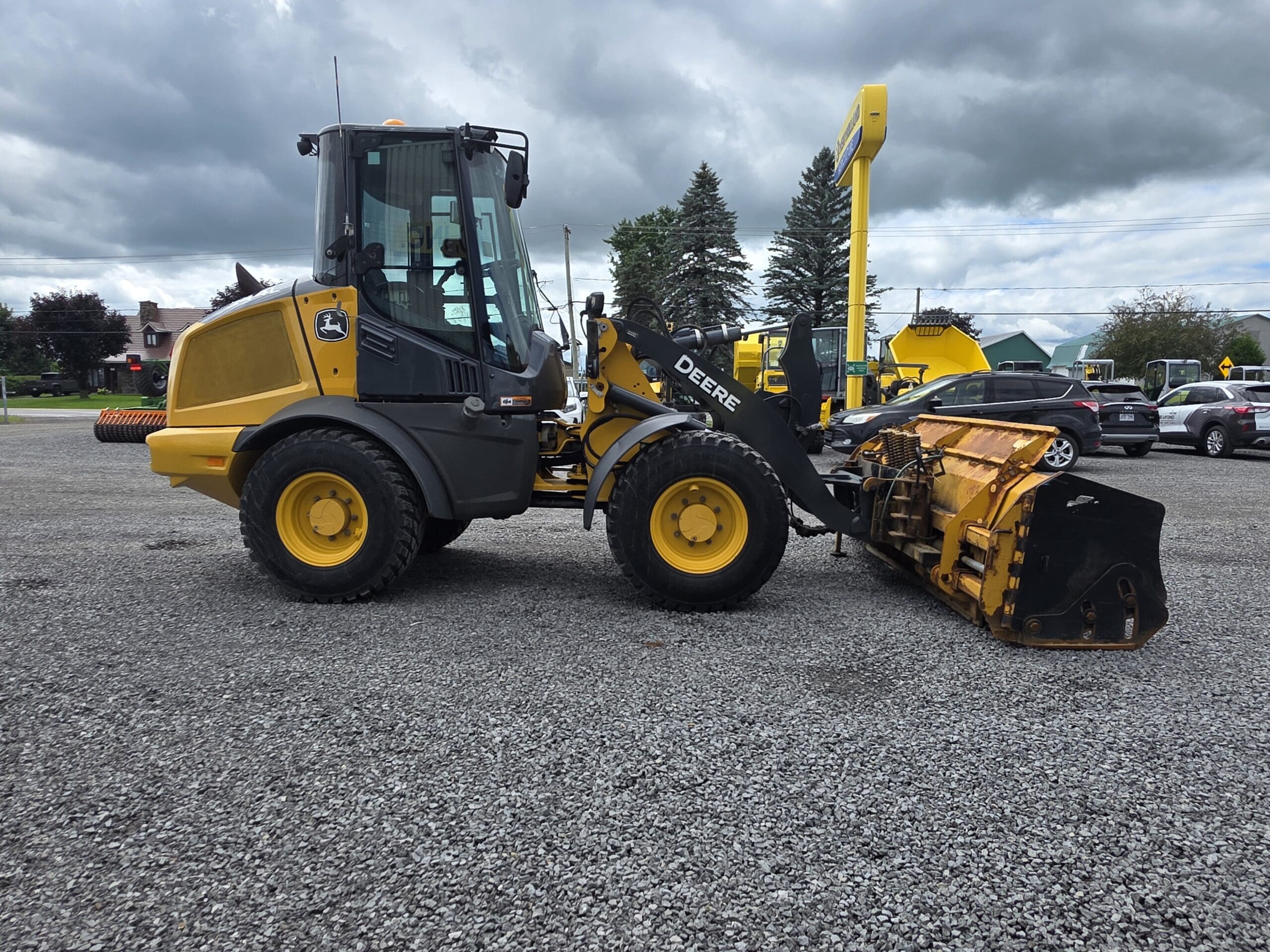 Une chargeuse frontale John Deere jaune équipée d'un gros accessoire est garée sur un terrain en gravier, avec d'autres véhicules et des arbres en arrière-plan, sous un ciel nuageux.