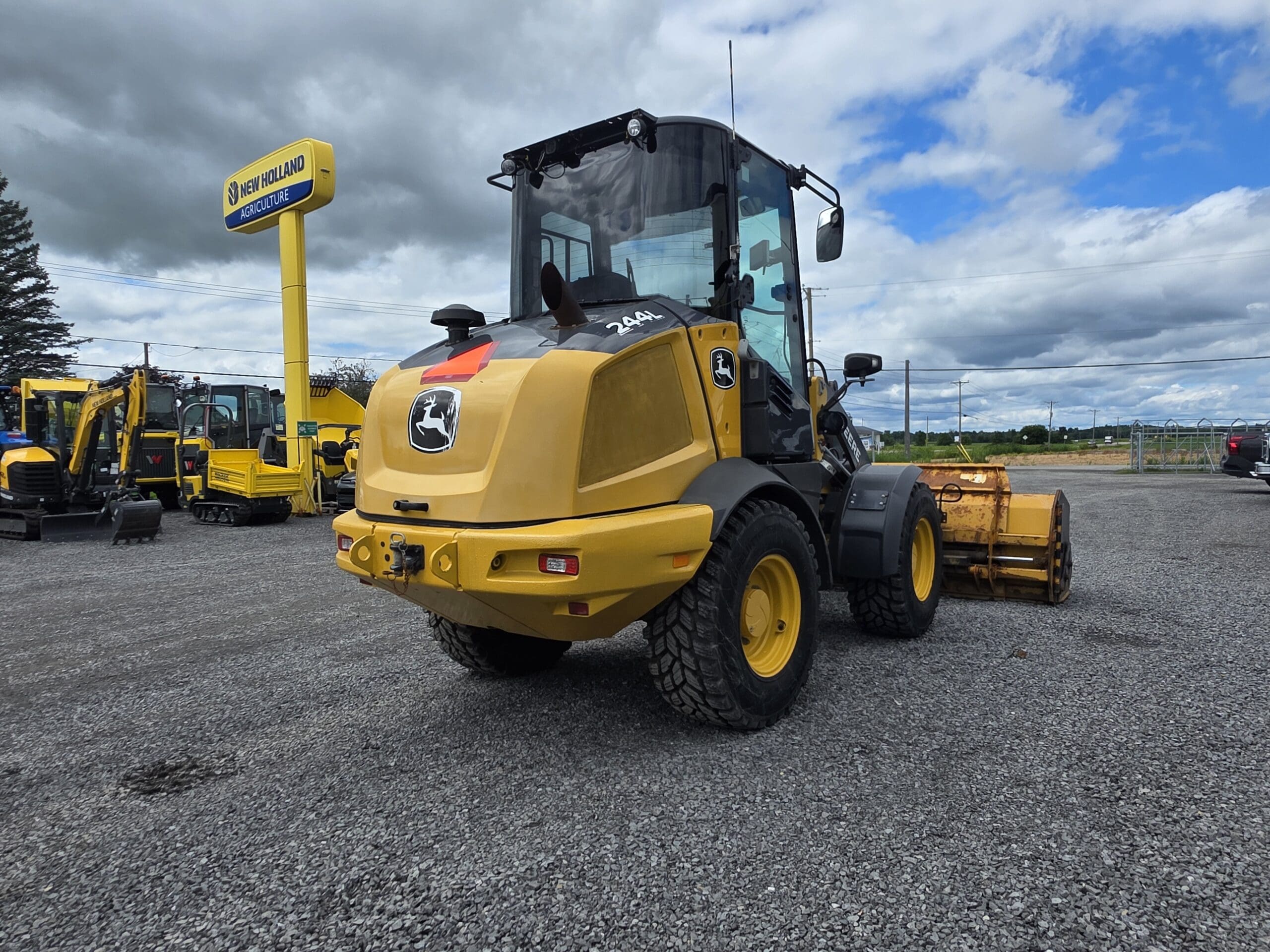 Une chargeuse John Deere jaune équipée d'un rouleau compresseur est garée sur du gravier près d'un panneau indiquant un concessionnaire New Holland, sous un ciel nuageux. On aperçoit des engins de chantier à l'arrière-plan.