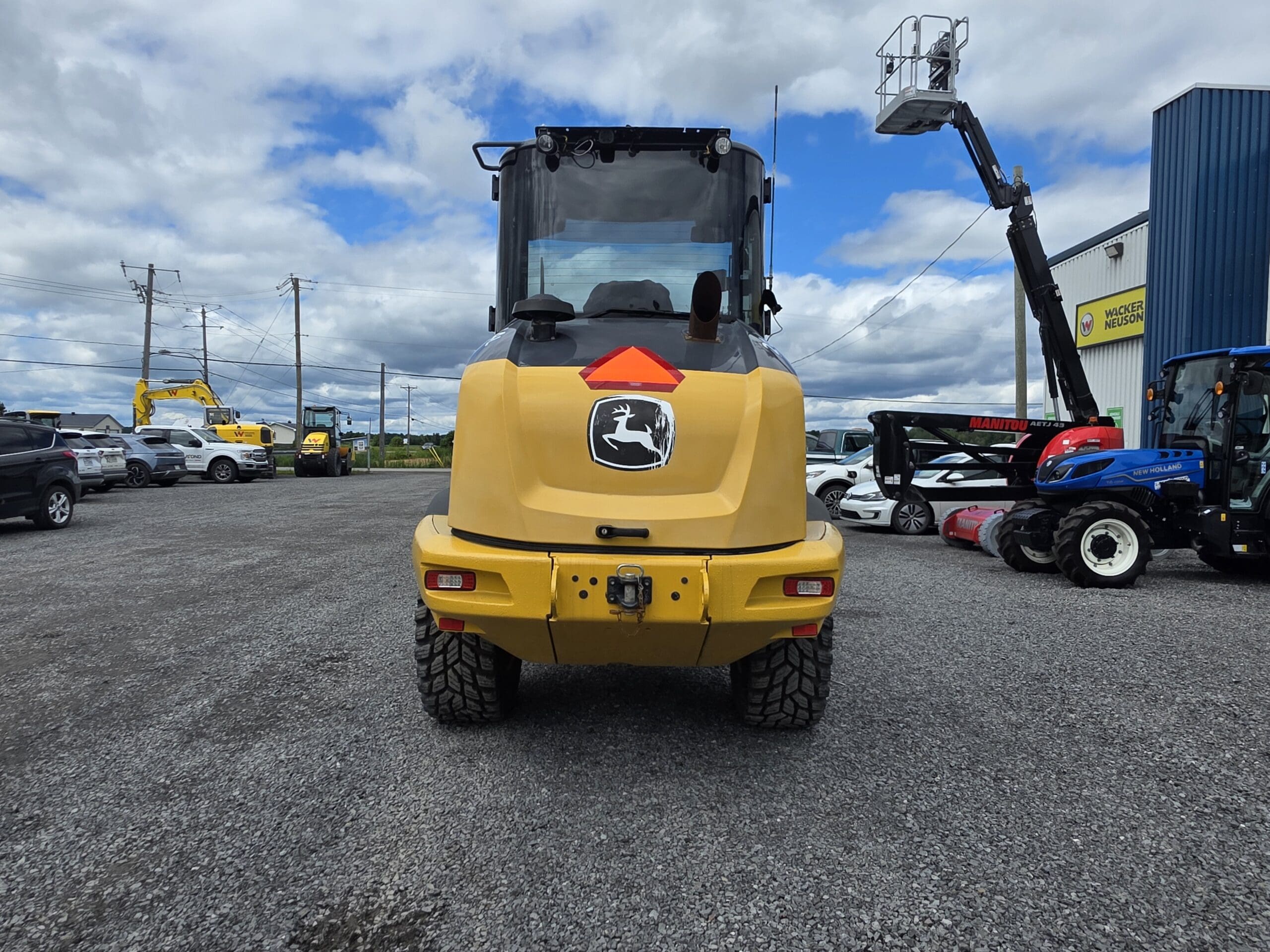 Un engin de chantier John Deere jaune est garé sur un terrain en gravier ; on le voit de dos, avec des voitures et du matériel visibles à l'arrière-plan, sous un ciel partiellement nuageux.