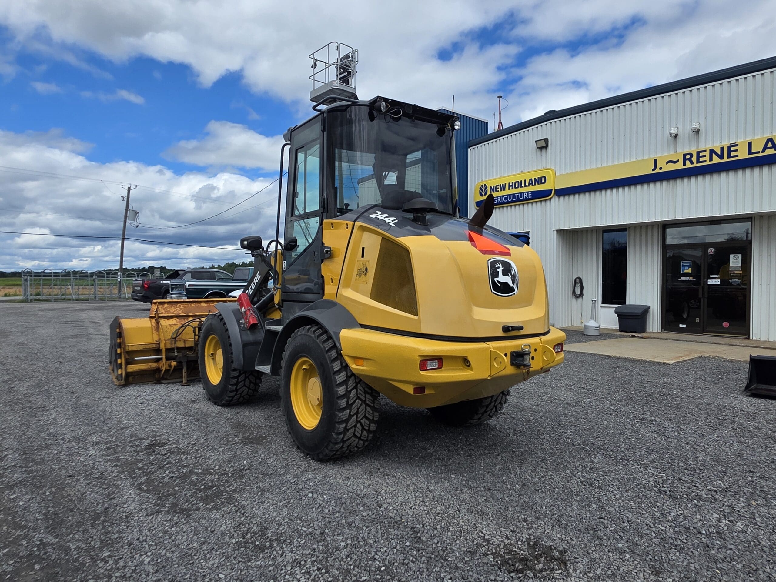 Une chargeuse frontale John Deere jaune, équipée de gros pneus, est garée sur du gravier devant un bâtiment arborant une enseigne « New Holland », sous un ciel partiellement nuageux.