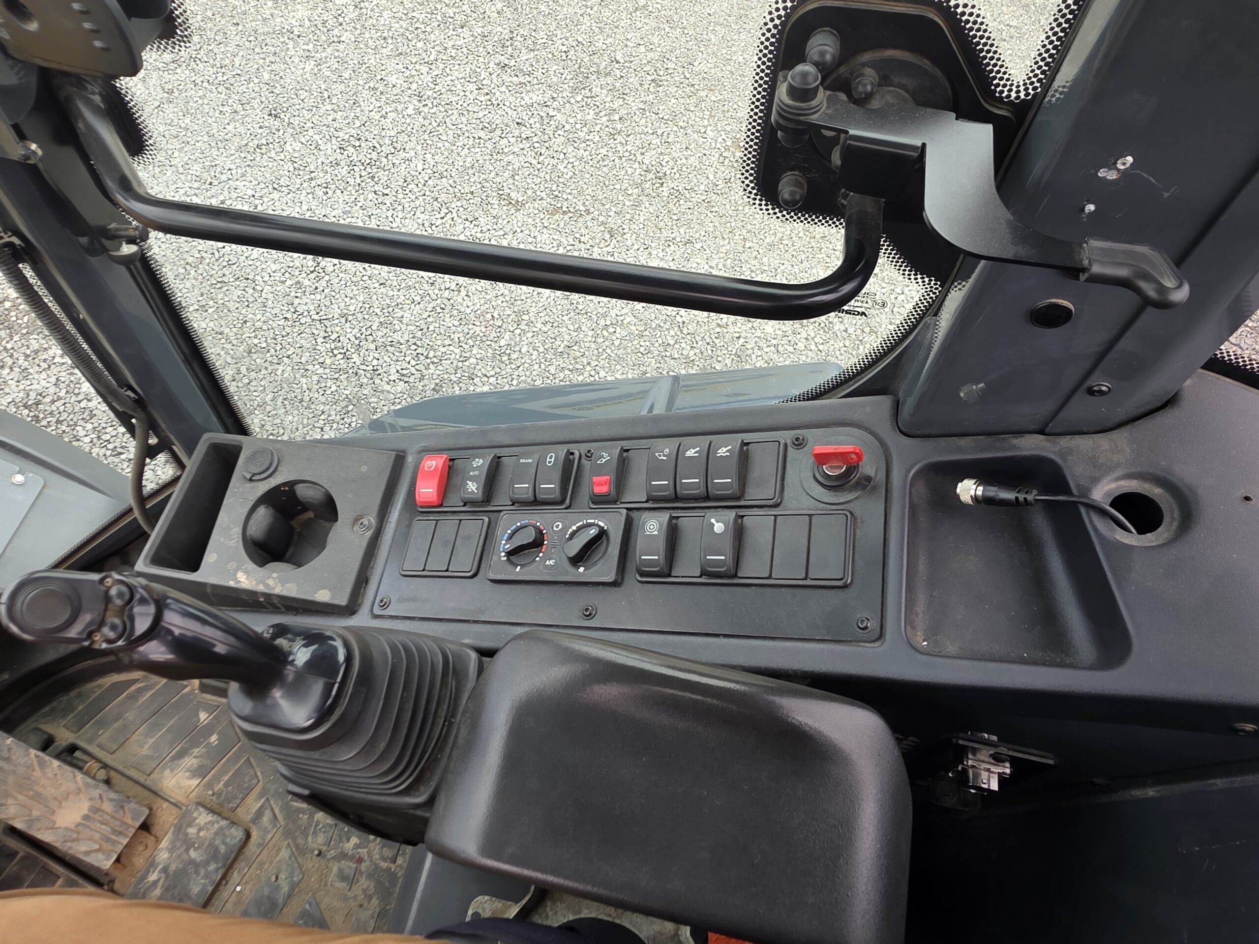Gros plan sur le tableau de bord d'un véhicule, comportant divers interrupteurs, boutons et un joystick. On aperçoit également un porte-gobelet et une vitre, à travers laquelle on distingue le sol à l'extérieur.