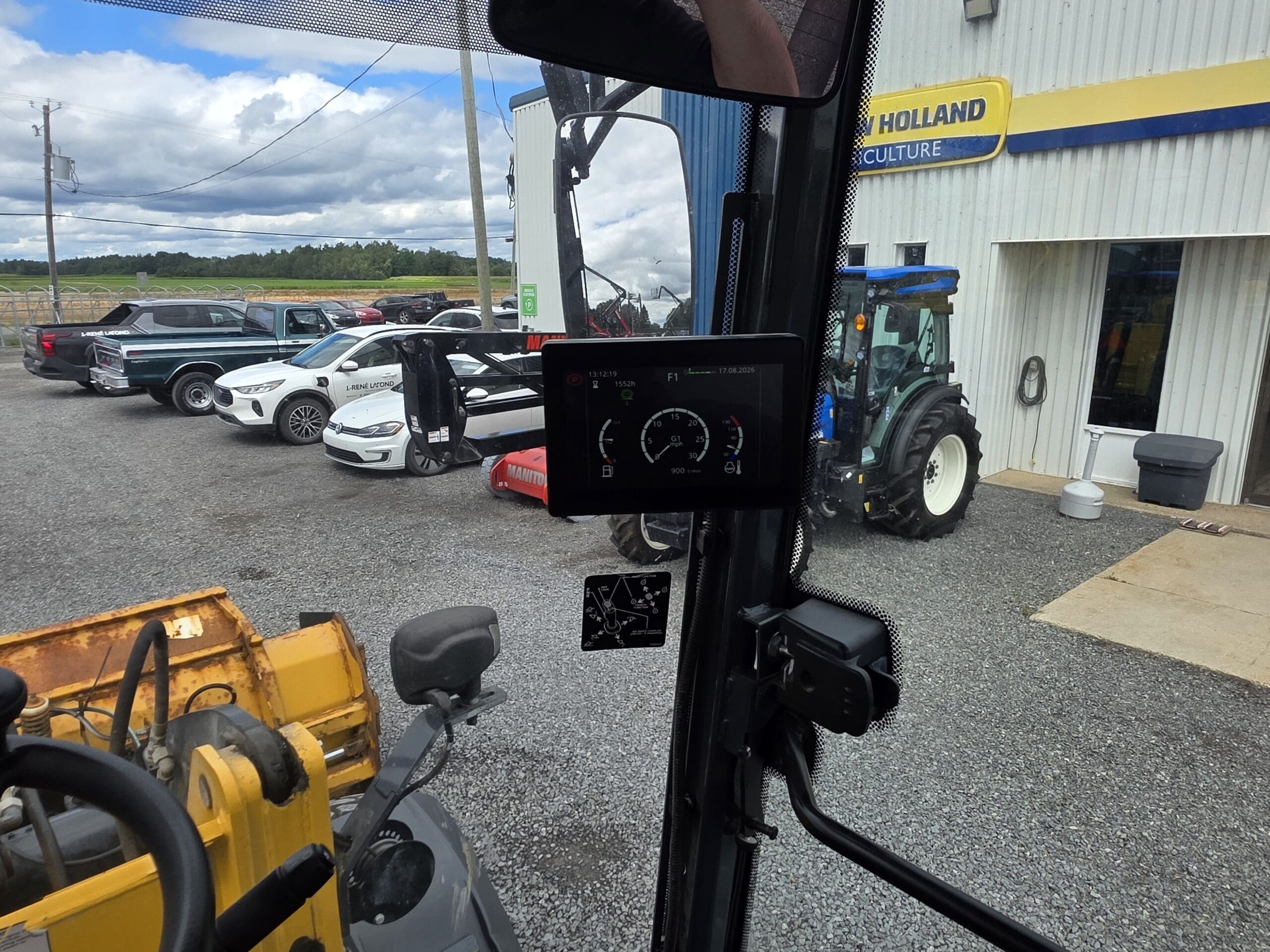 Vue depuis l'intérieur d'un véhicule montrant l'écran numérique du tableau de bord, avec, à l'arrière-plan, des voitures garées, un tracteur et un bâtiment de New Holland Agriculture sous un ciel nuageux.