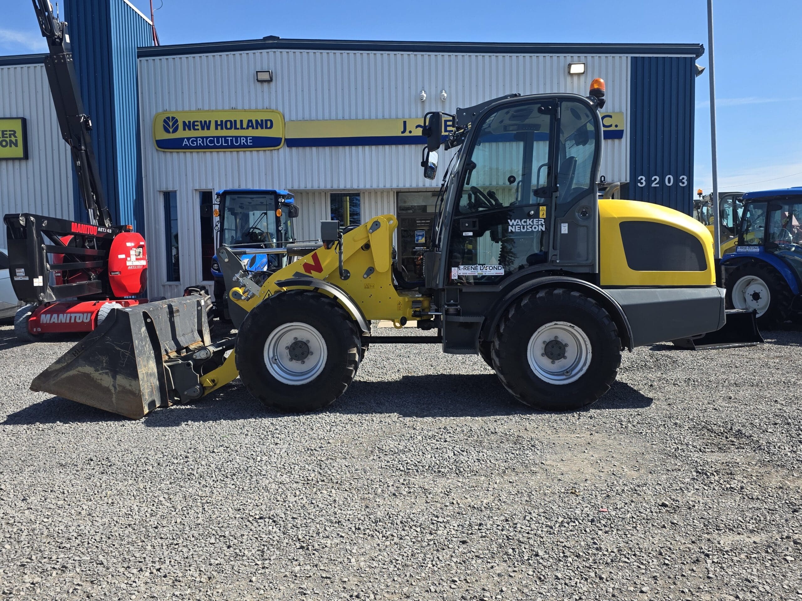 Une chargeuse sur pneus compacte jaune et noire est garée sur du gravier devant un bâtiment de New Holland Agriculture ; on aperçoit d'autres machines et tracteurs à l'arrière-plan.