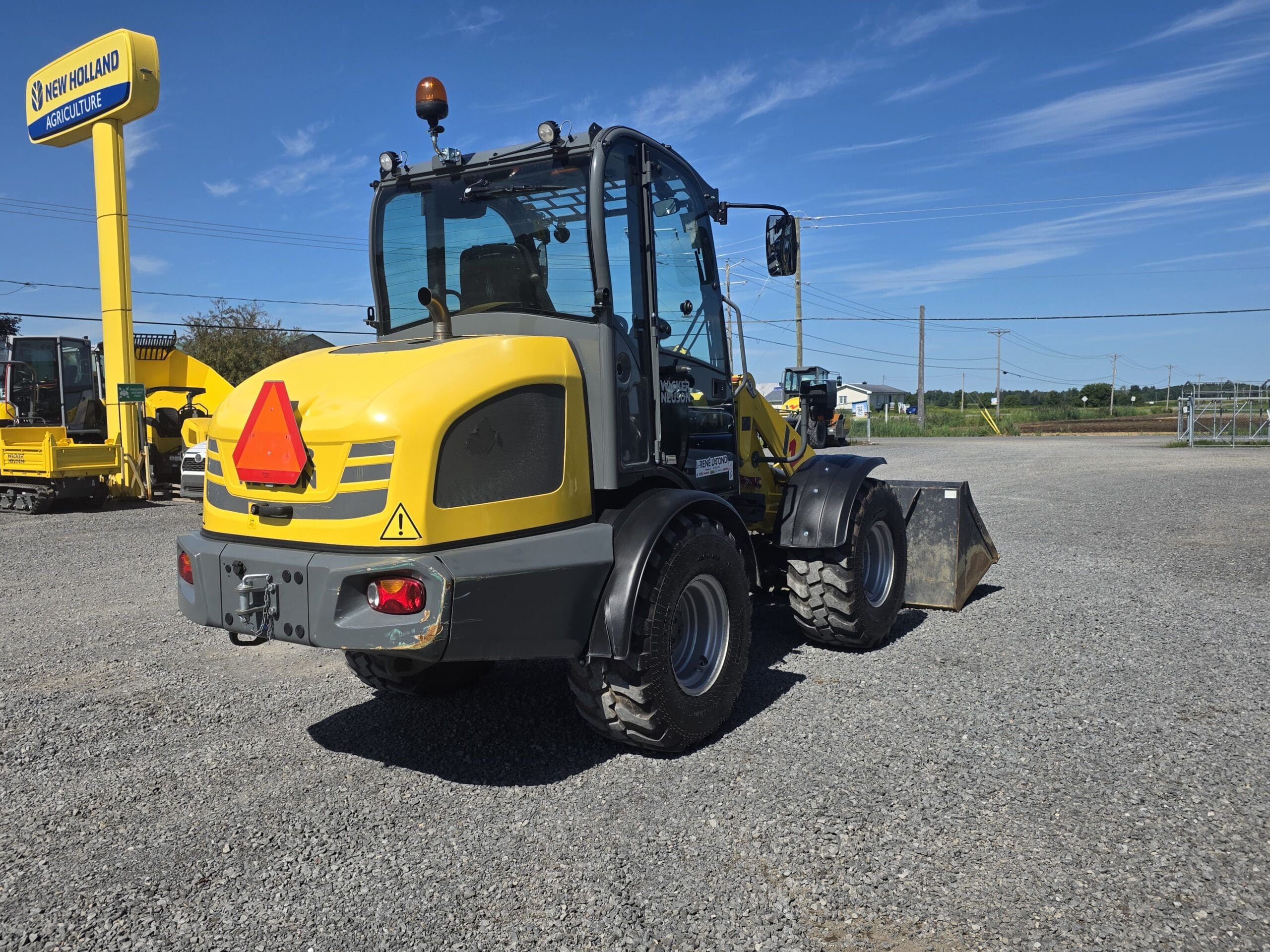 Un chargeur frontal jaune est garé sur du gravier dans un parking, sous un ciel bleu limpide ; il porte à l'arrière un triangle rouge signalant un véhicule lent, et on aperçoit en arrière-plan l'enseigne « New Holland Agriculture ».