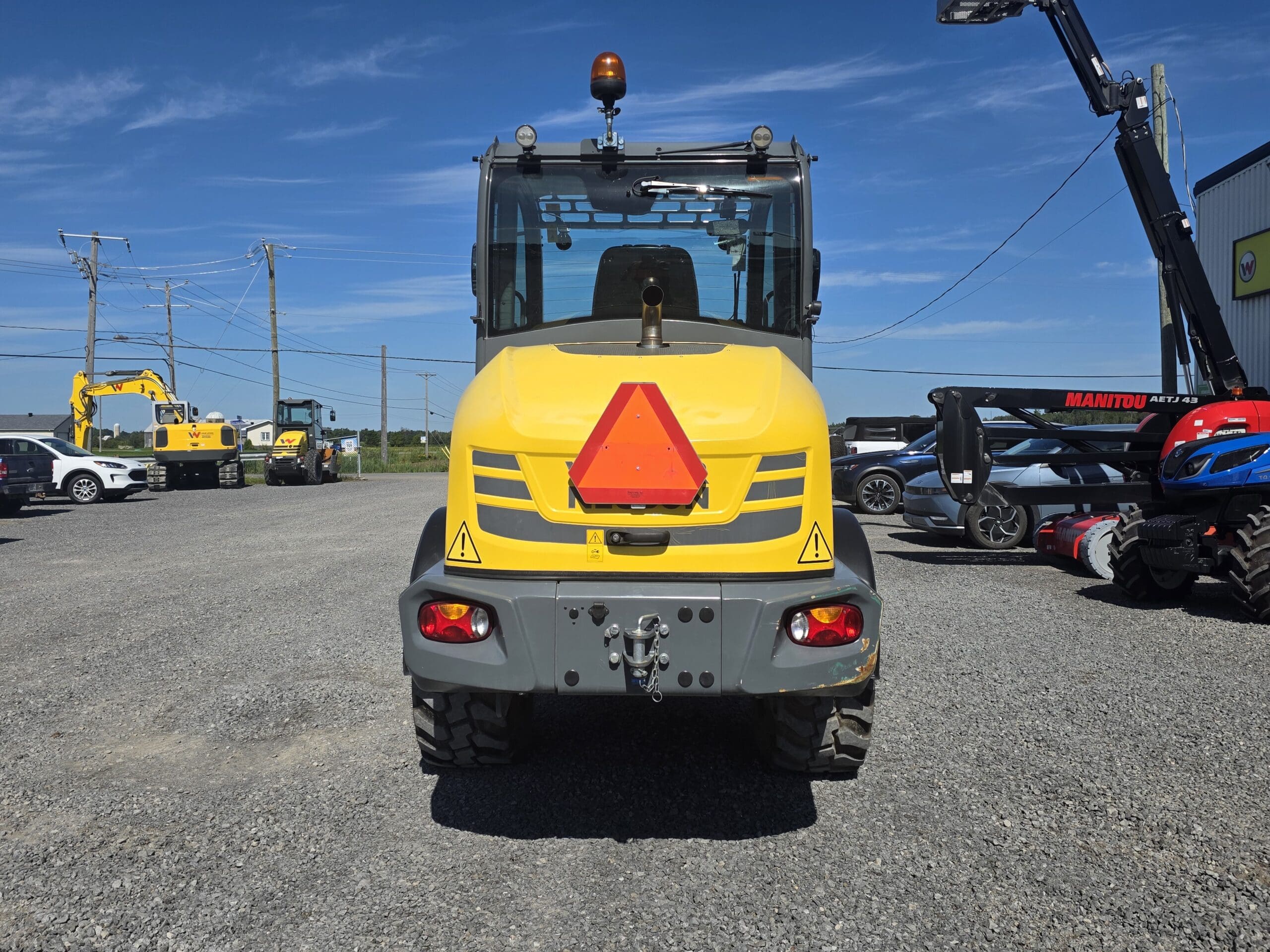 Un engin de chantier jaune est garé sur un terrain en gravier ; il est photographié de dos. D'autres engins, des voitures et des lignes électriques sont visibles à l'arrière-plan, sous un ciel bleu limpide.