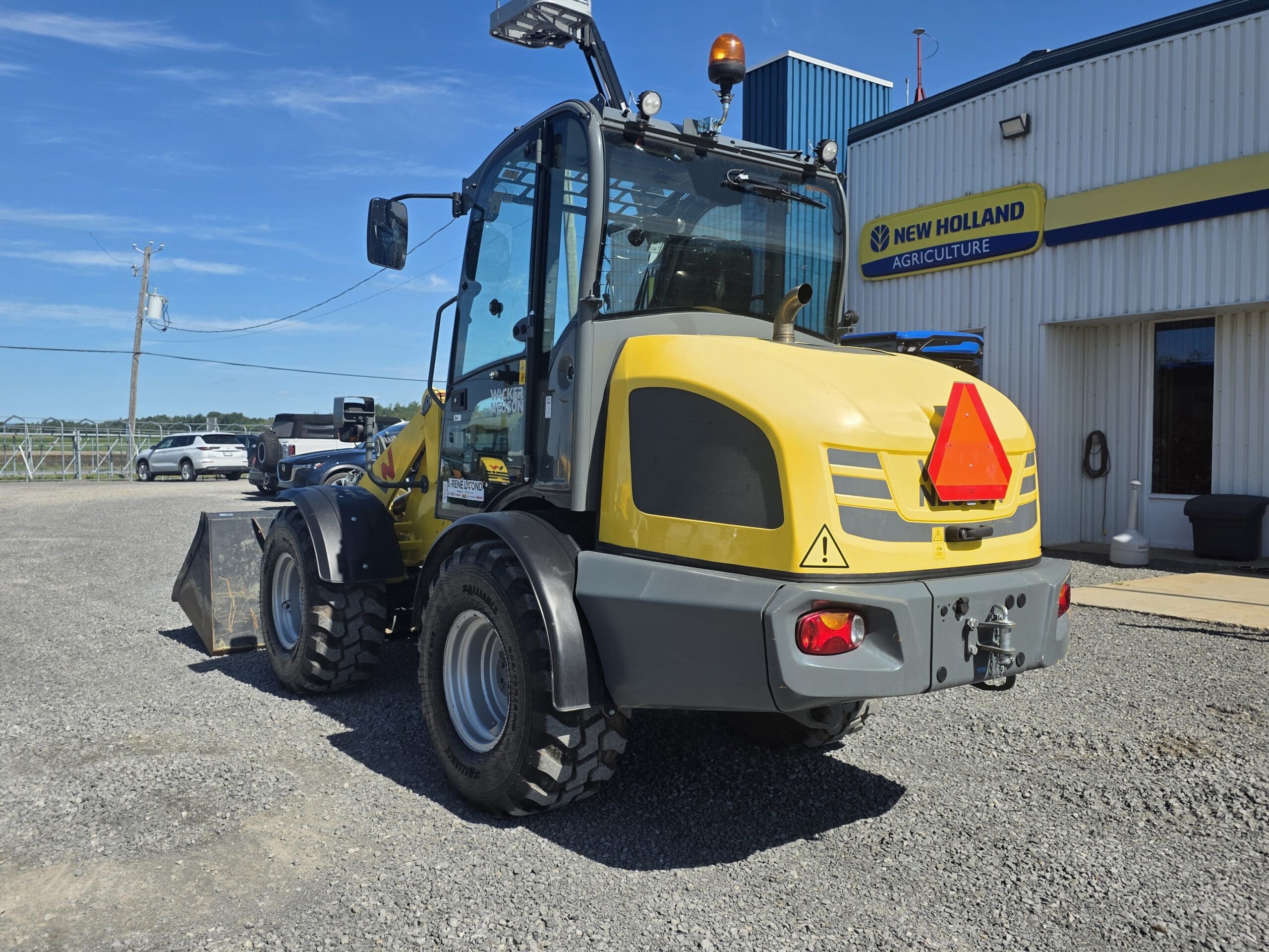 Une chargeuse frontale jaune et grise est garée sur du gravier près d'un bâtiment arborant une enseigne « New Holland Agriculture ». Le ciel est dégagé et bleu.