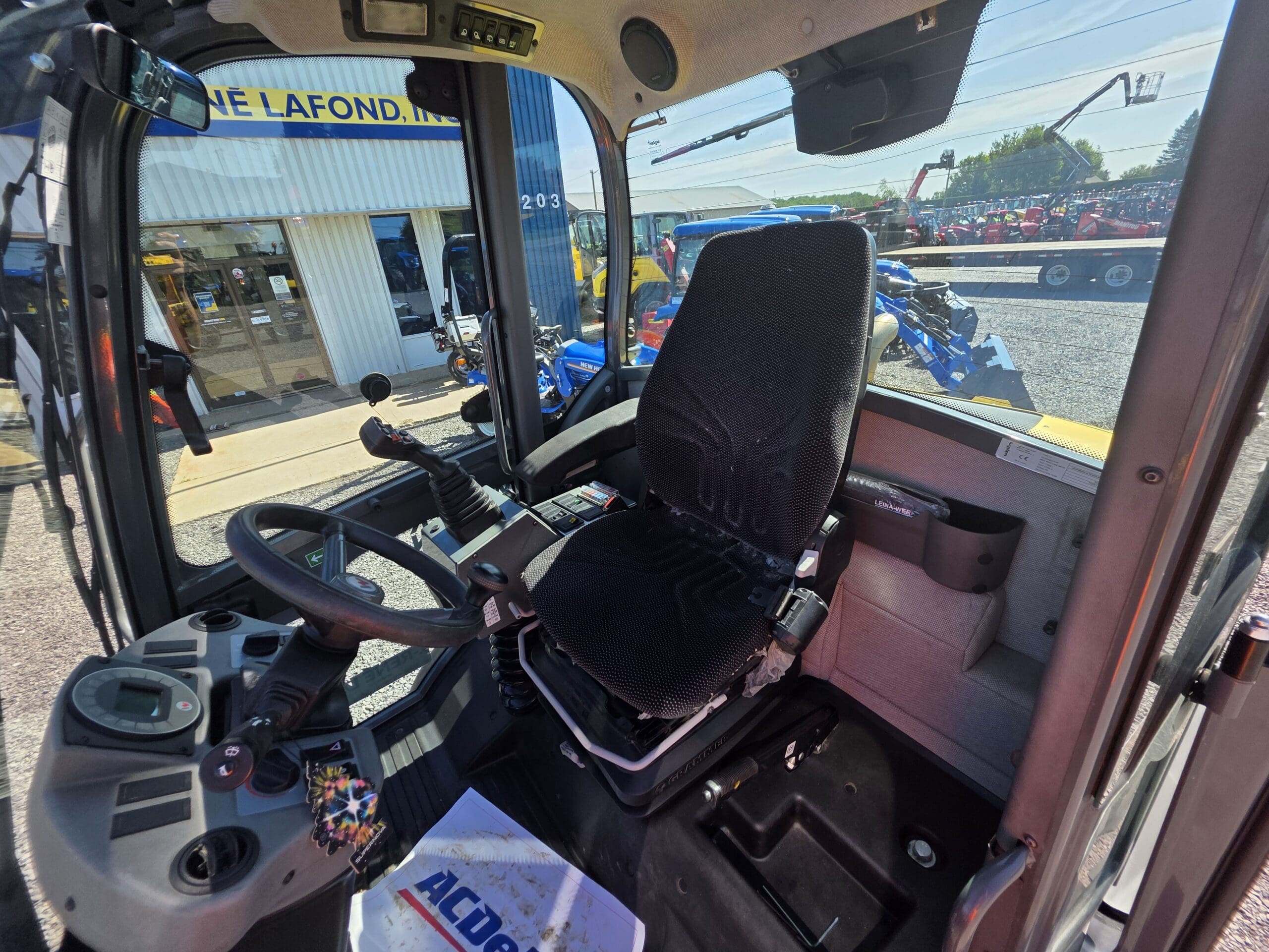 Vue intérieure de la cabine d'un tracteur montrant le siège du conducteur, le volant et le tableau de bord, ainsi qu'une vue extérieure d'un concessionnaire où l'on aperçoit des tracteurs et du matériel à travers les vitres.