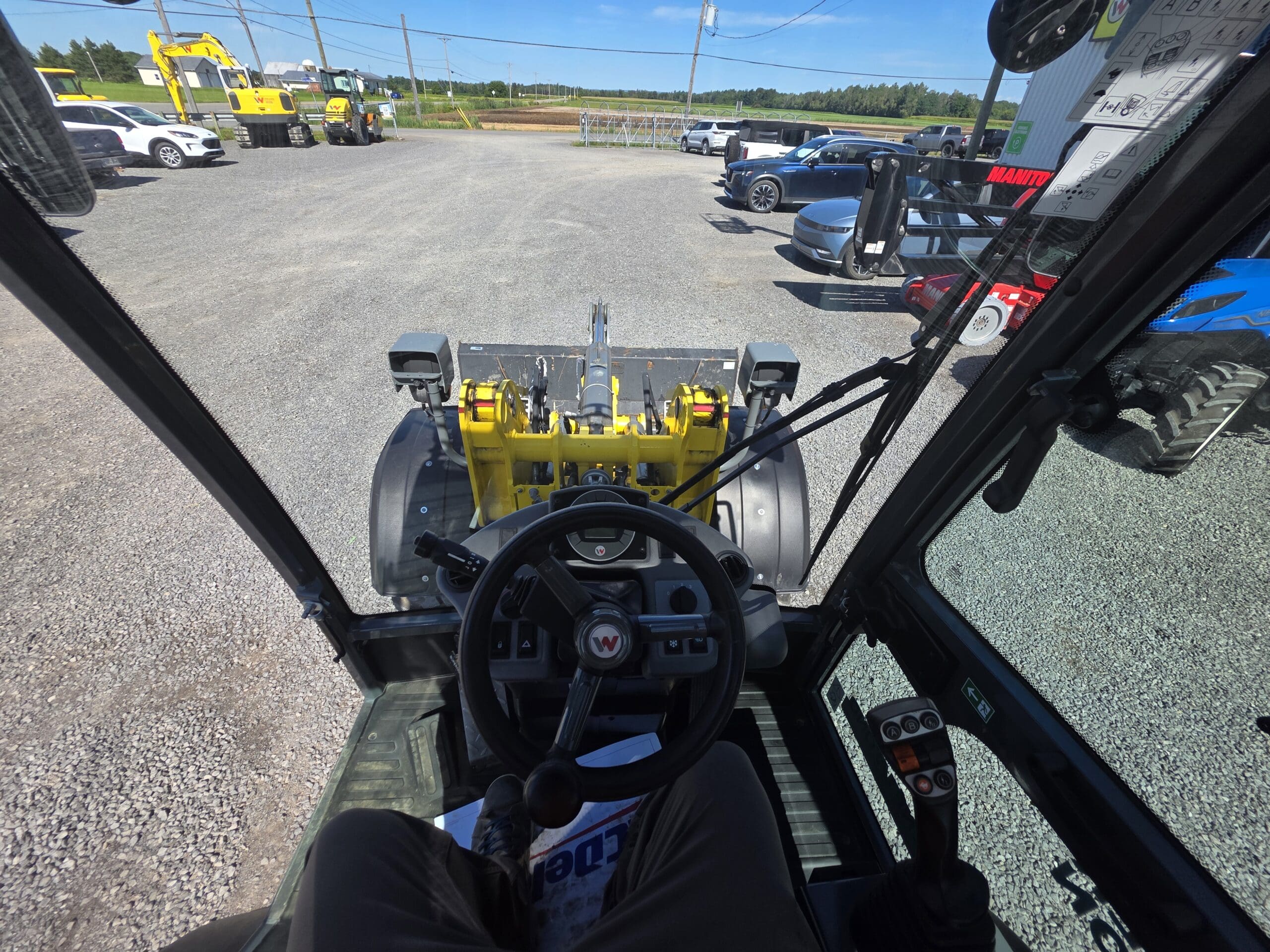 Vue depuis l'intérieur de la cabine d'un engin de chantier, montrant le volant, les commandes et un godet jaune, avec divers véhicules et engins visibles à l'extérieur sur un terrain en gravier.