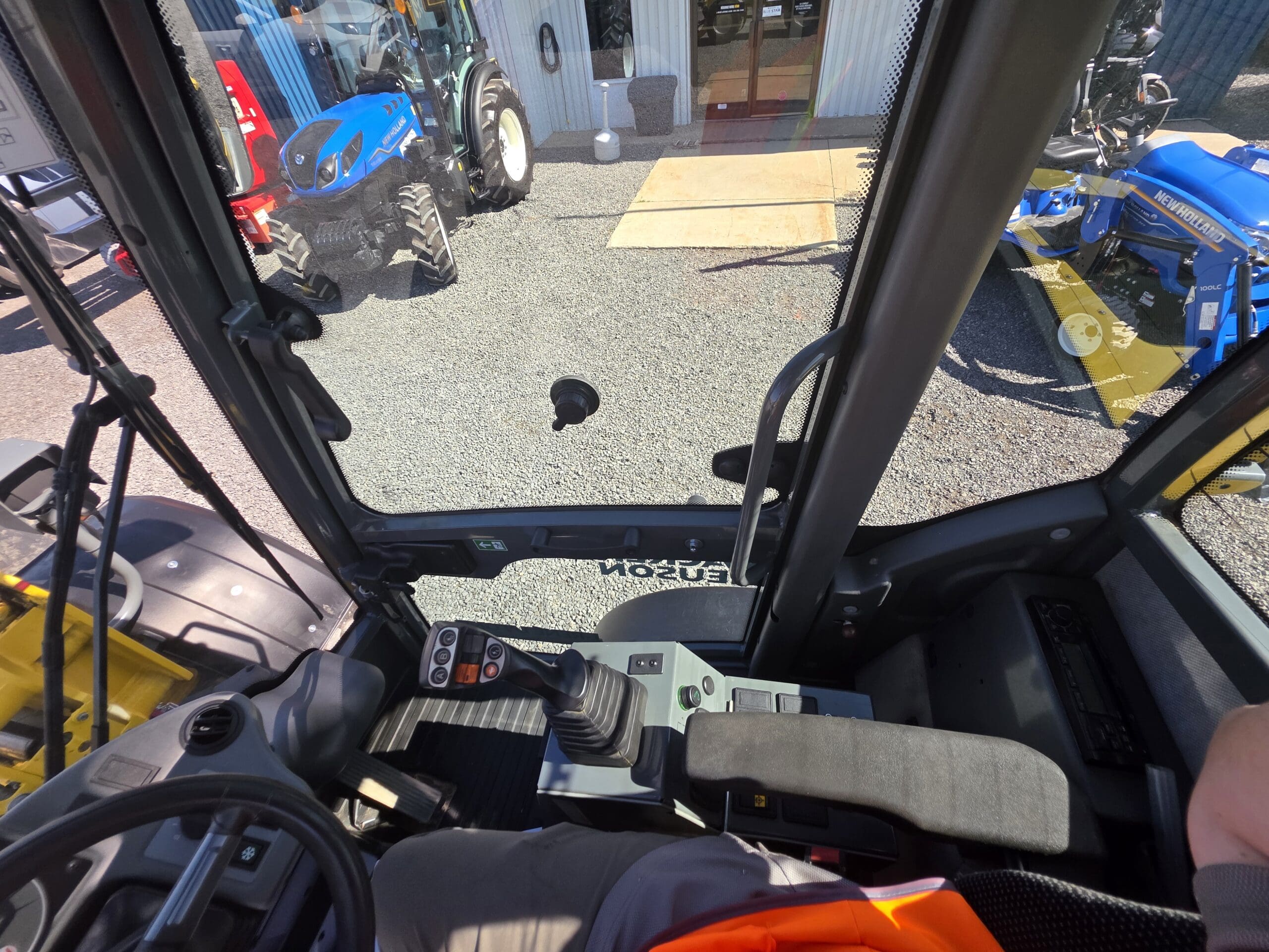Vue depuis l'intérieur de la cabine d'un engin de chantier montrant le tableau de bord, le joystick, le siège et le bras de l'opérateur. À l'extérieur de la cabine, on aperçoit des tracteurs et du gravier en plein soleil.