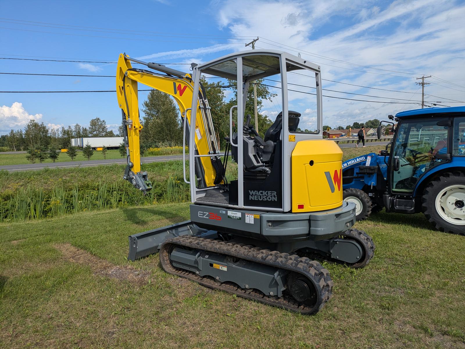 Wacker Neuson EZ36 vu de côté, bras articulé relevé, tracteur bleu en arrière-plan