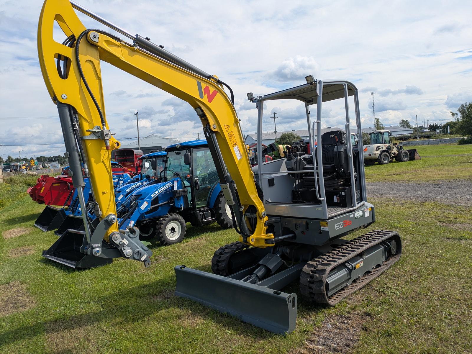 Wacker Neuson EZ36 vu de trois quarts avant-gauche, bras articulé étendu avec godet, tracteur bleu en arrière-plan