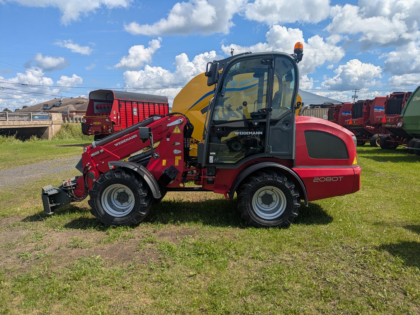 Weidemann 2080T vu de côté, bras télescopique relevé, autres équipements agricoles en arrière-plan