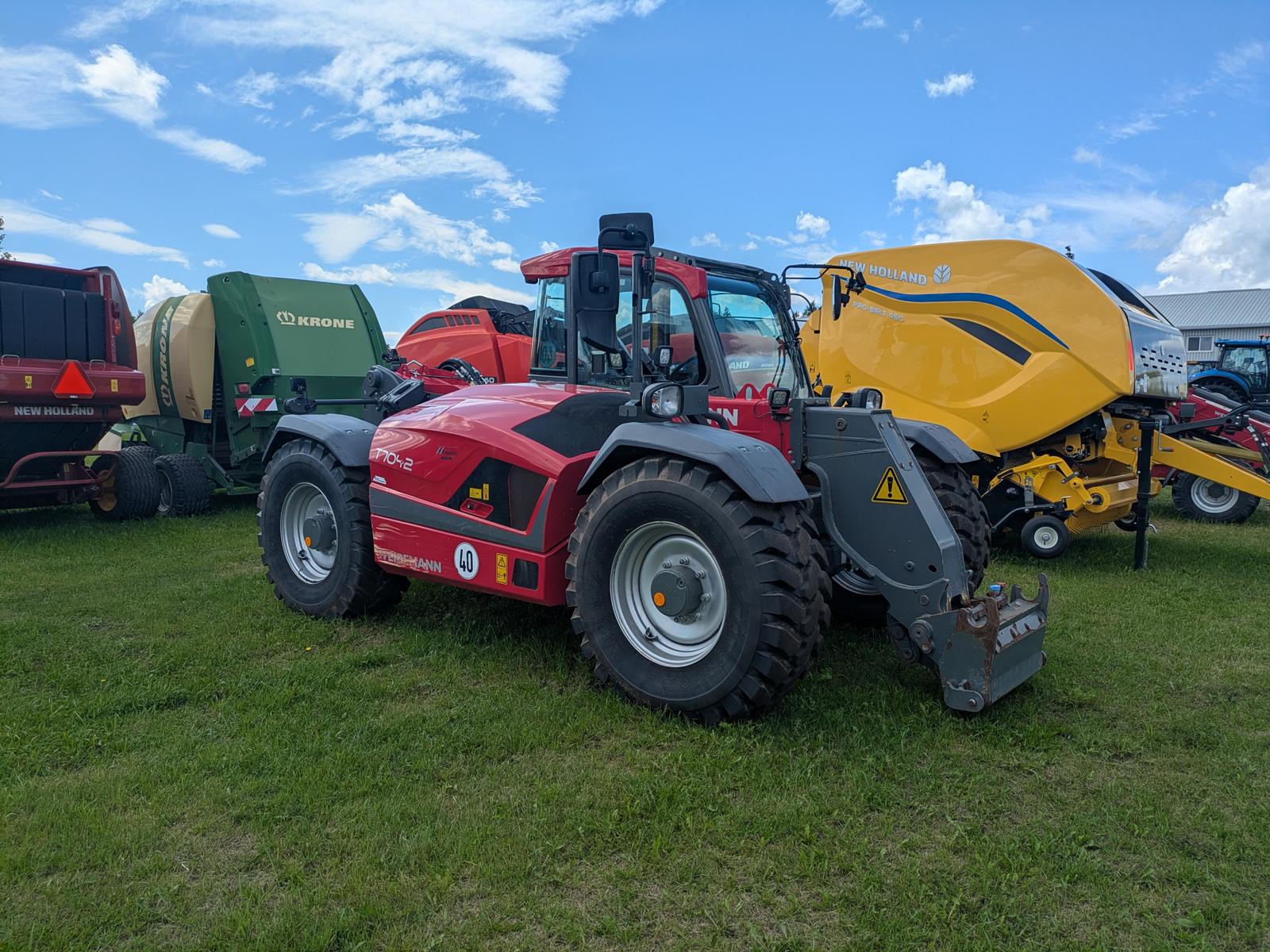 Weidemann 2080T vu de trois quarts avant avec une enrouleuse à foin jaune attelée, autres machines agricoles en arrière-plan