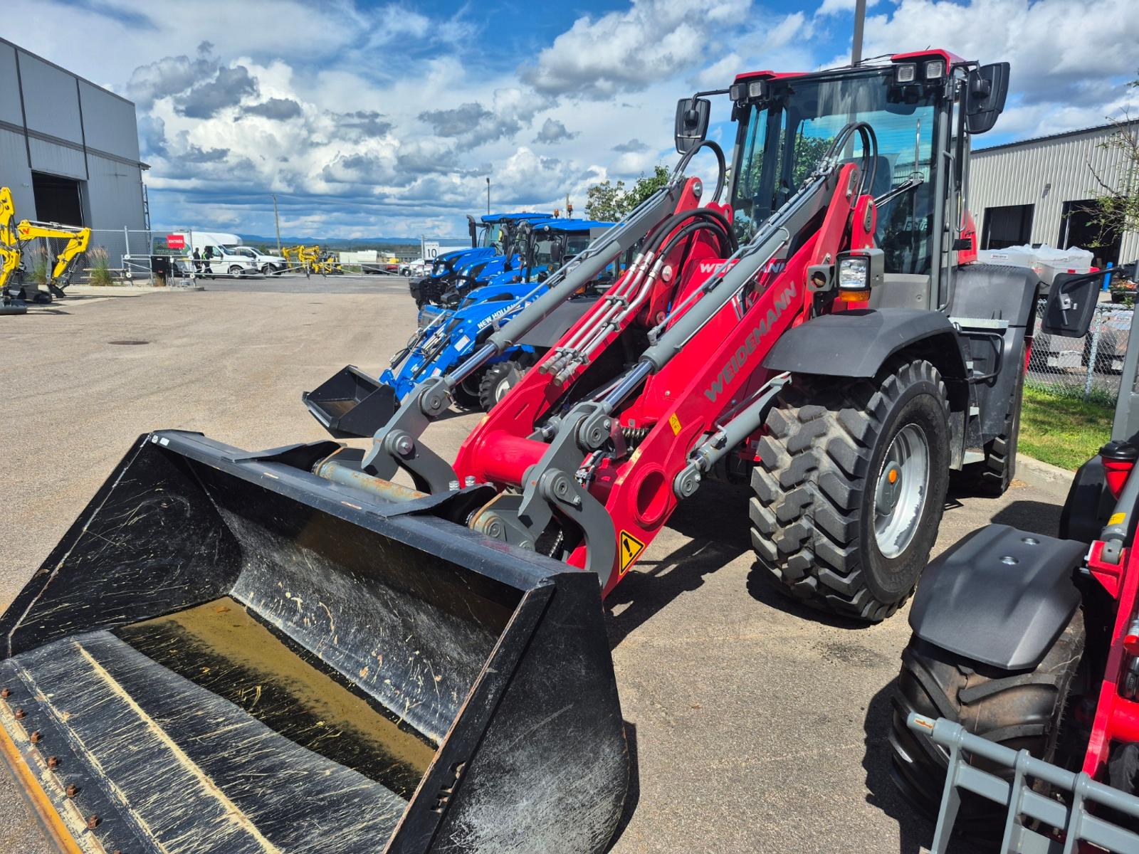 Weidemann 9080 vu de trois quarts avant, godet levé, autres machines stationnées en arrière-plan