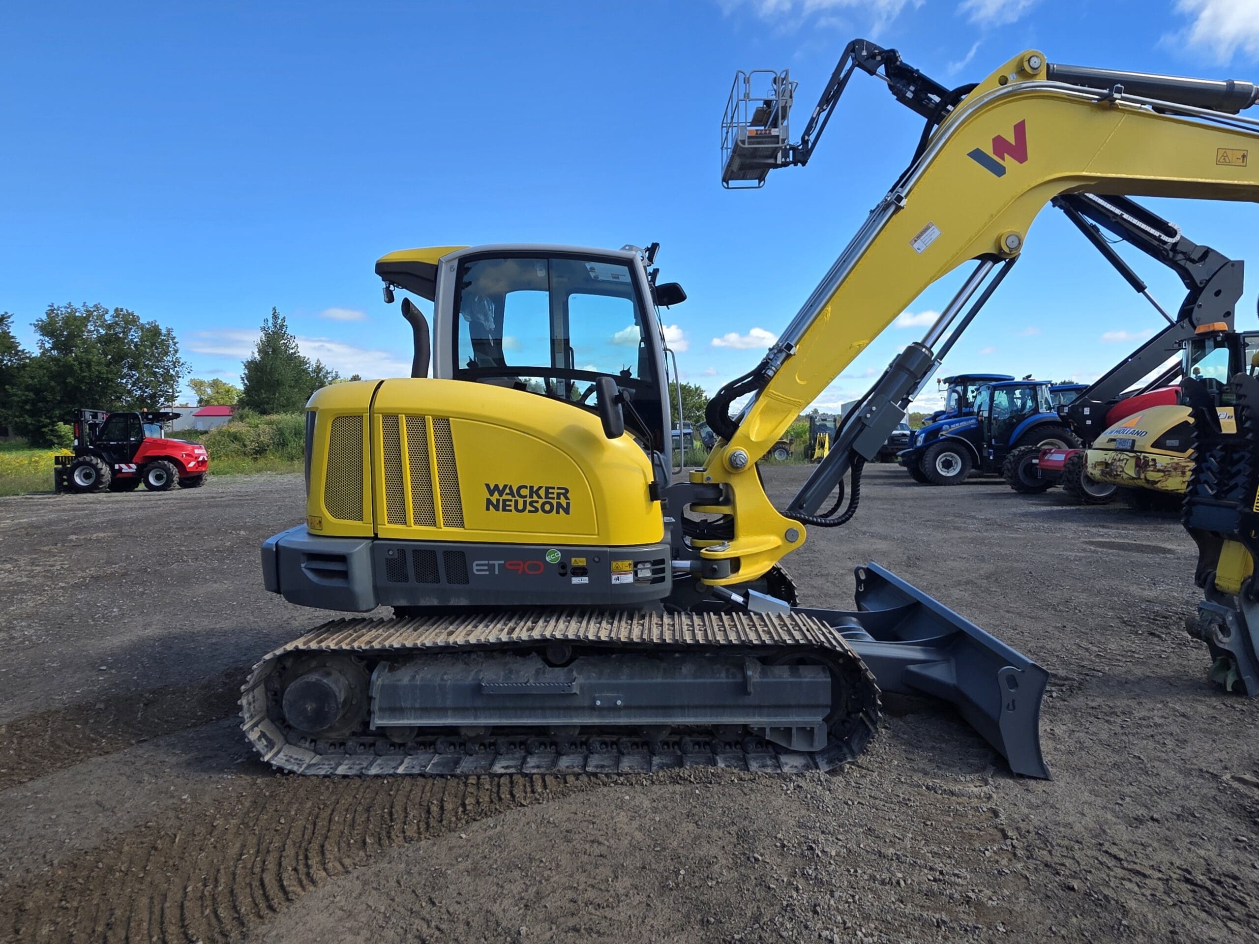 Une pelleteuse sur chenilles Wacker Neuson ET90 de couleur jaune est garée sur un terrain en gravier, avec d'autres engins de chantier visibles à l'arrière-plan sous un ciel bleu.