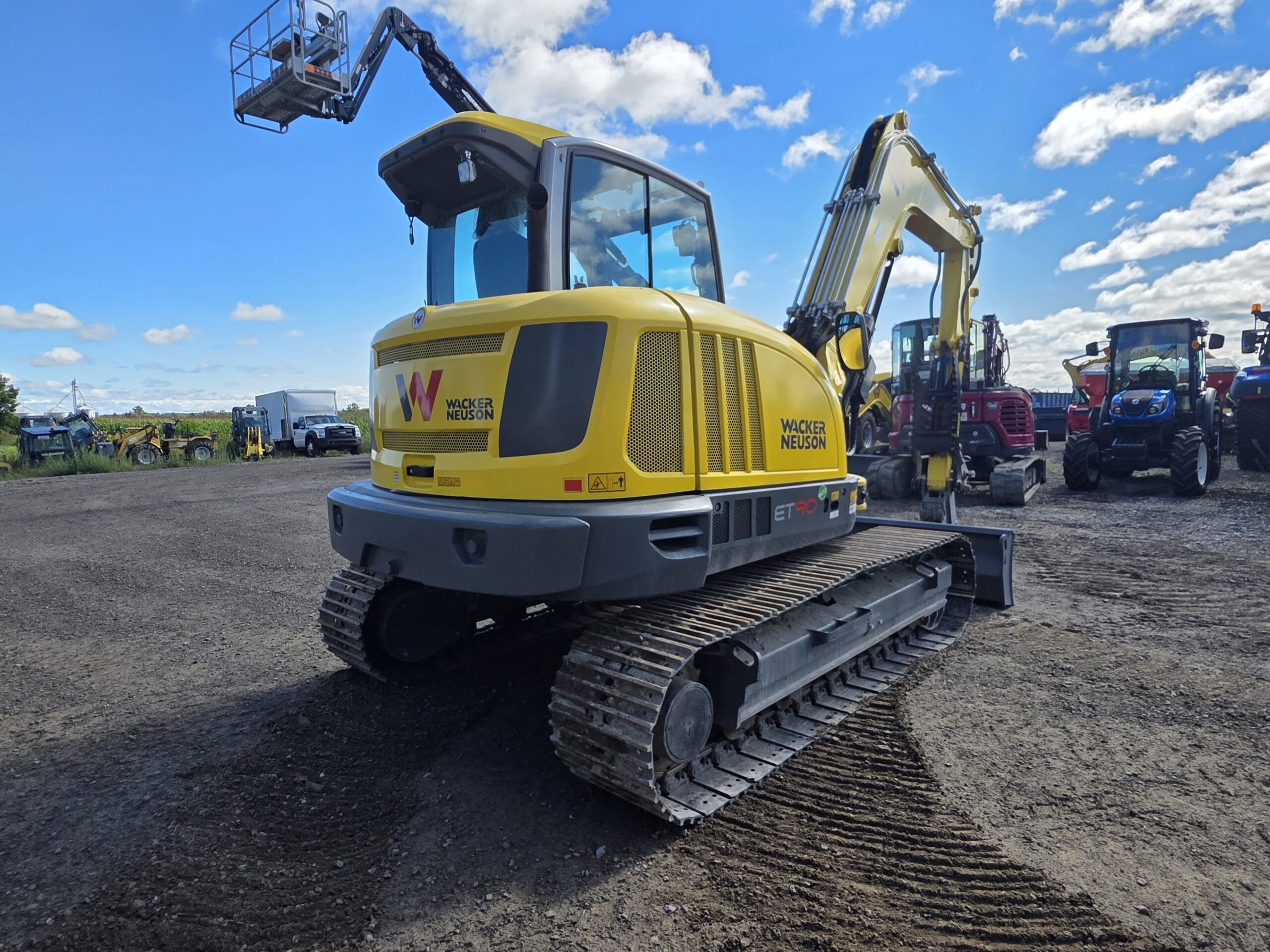 Une pelleteuse sur chenilles jaune de la marque Wacker Neuson est garée sur du gravier, dans un parking où se trouvent d'autres engins de chantier et véhicules, sous un ciel bleu partiellement nuageux.