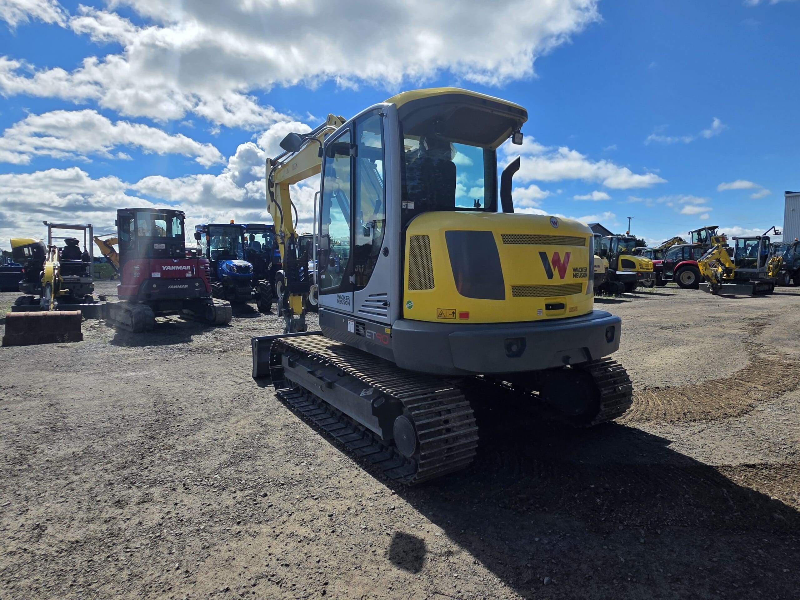 Une pelleteuse à chenilles jaune se trouve sur un terrain en gravier sous un ciel partiellement nuageux, entourée d'autres engins et équipements de chantier en arrière-plan.