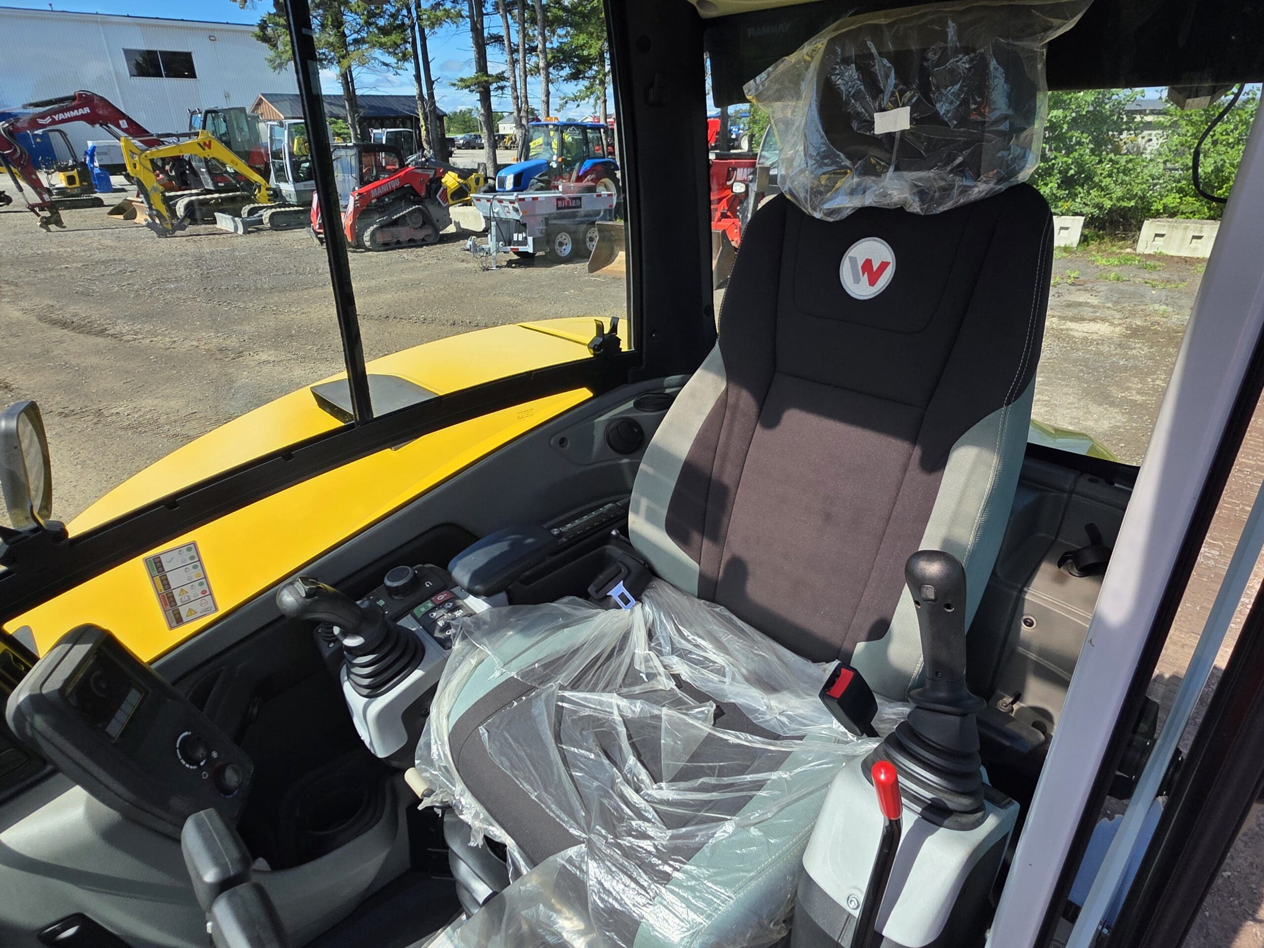 Siège conducteur et commandes à l'intérieur d'un engin de chantier, recouverts de plastique. À l'extérieur, on aperçoit du matériel et des véhicules de chantier à travers les vitres.