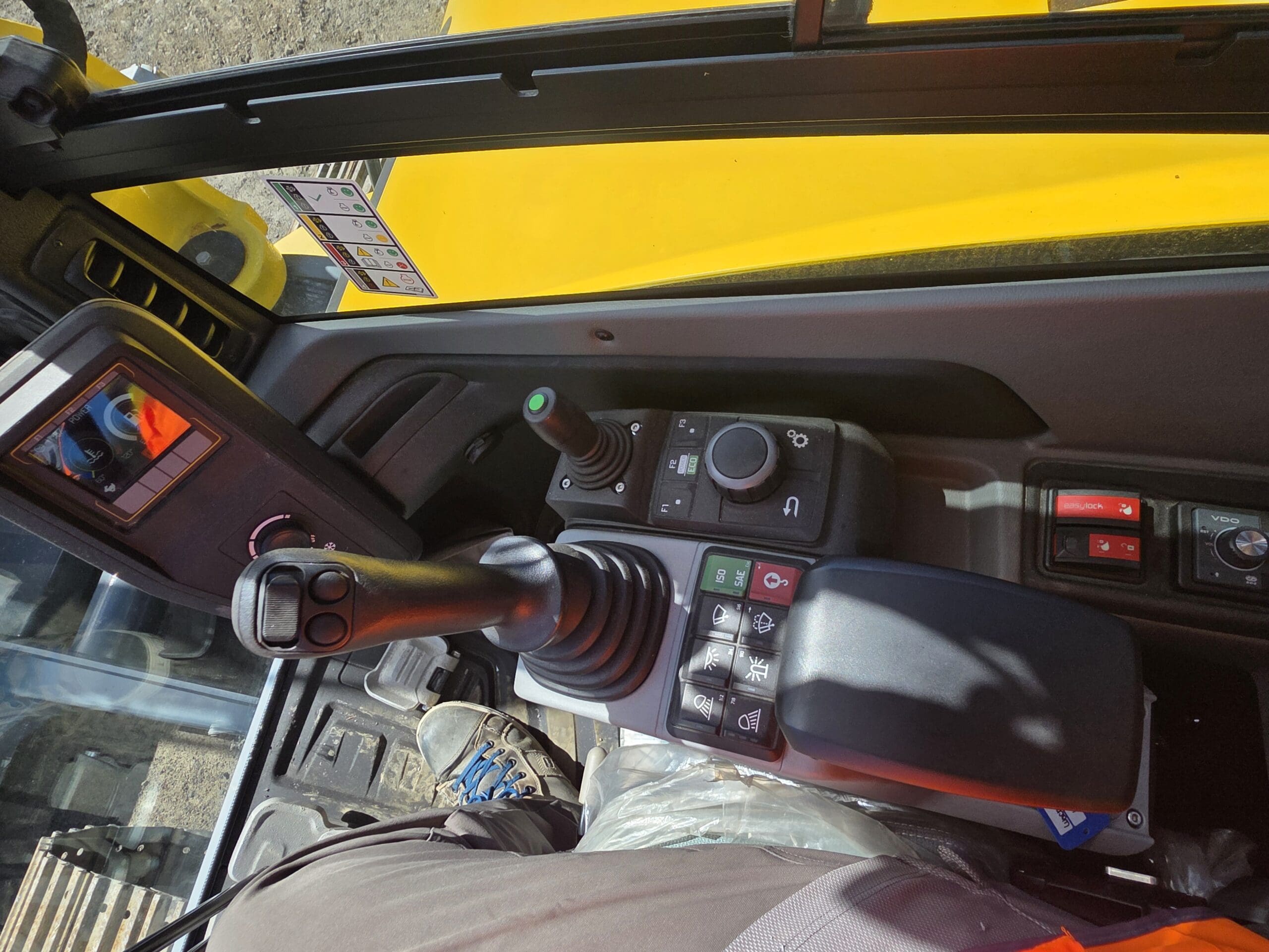Vue depuis l'intérieur de la cabine d'un engin de chantier, montrant les leviers de commande, les boutons et une partie de la jambe du conducteur. La lumière du soleil met en valeur la carrosserie jaune visible à travers la vitre.