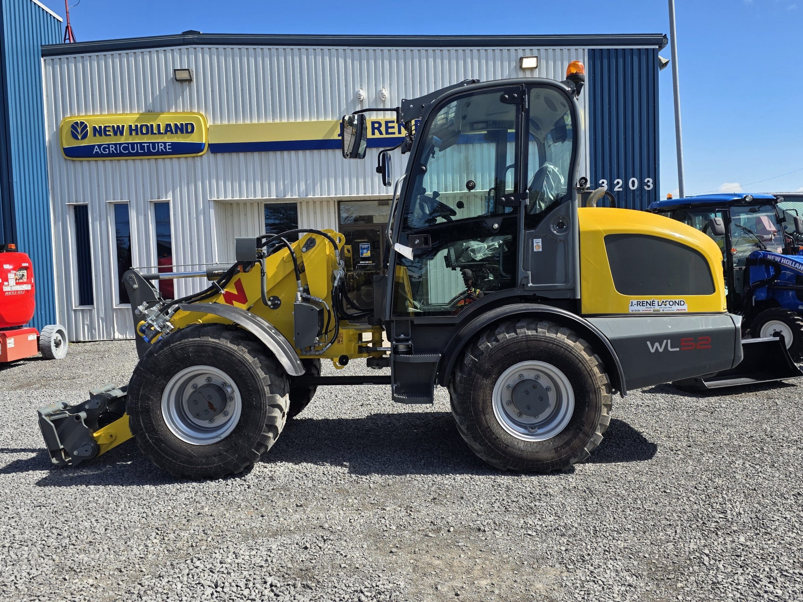 Une chargeuse sur pneus WL52 jaune et noire est garée sur du gravier devant un bâtiment de New Holland Agriculture, sous un ciel bleu limpide.