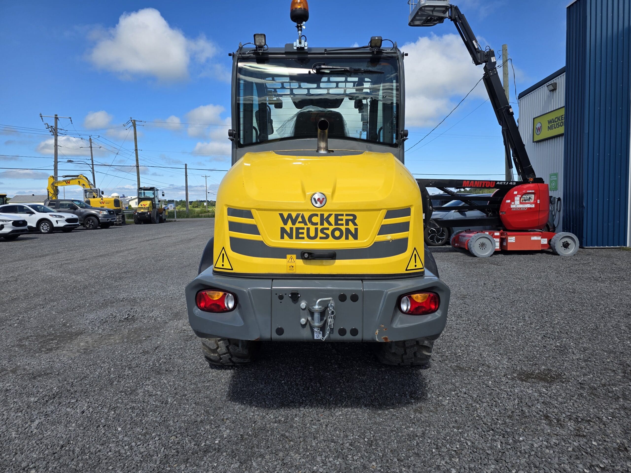 Un engin de chantier jaune de la marque Wacker Neuson est garé sur du gravier, vu de l'arrière. D'autres engins et un bâtiment sont visibles à l'arrière-plan, sous un ciel bleu parsemé de nuages.