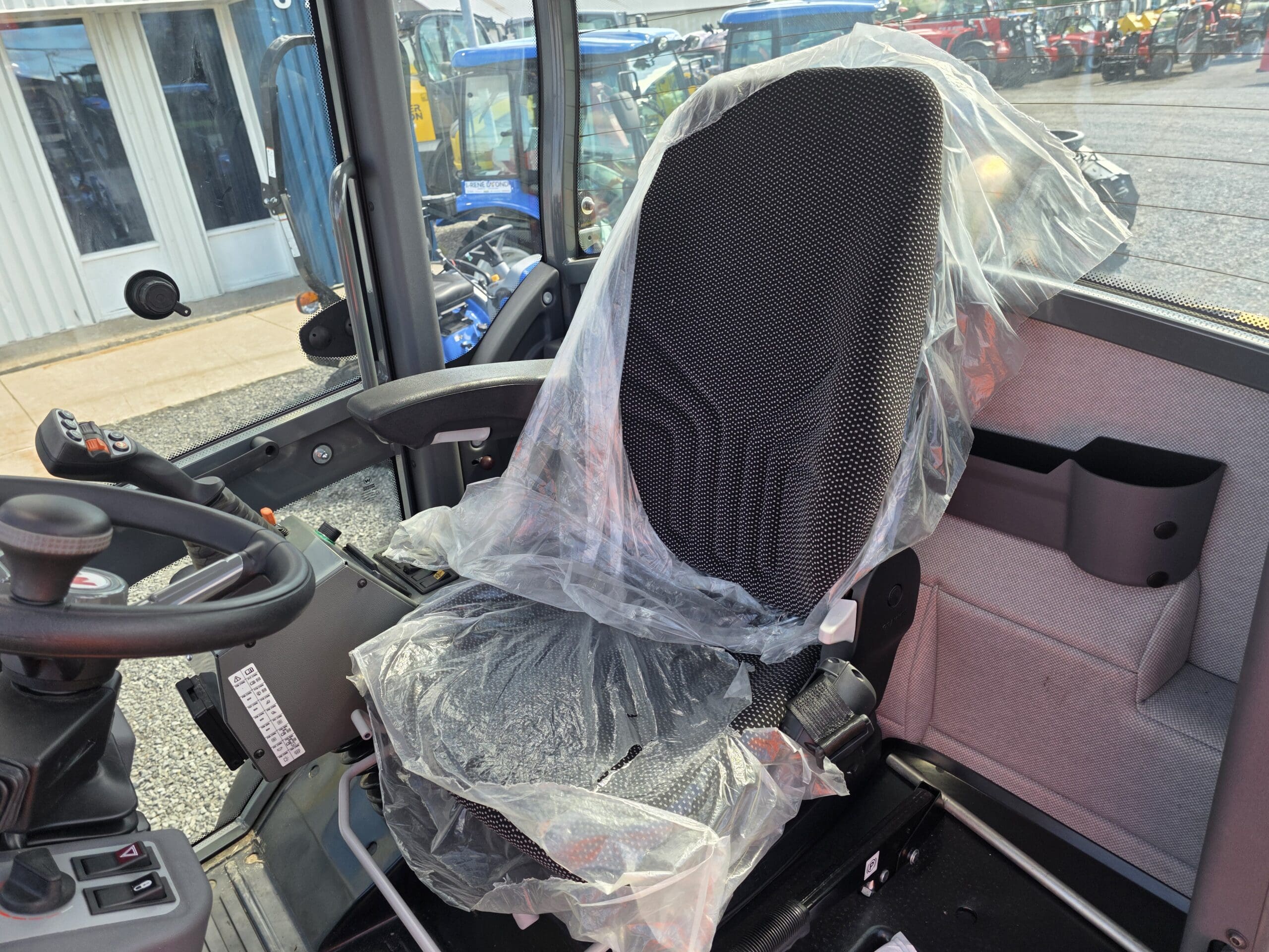 À l'intérieur de la cabine d'un véhicule se trouve un siège conducteur noir recouvert d'un film plastique de protection, avec des leviers de commande à proximité et, à l'arrière-plan, des vitres donnant sur des équipements extérieurs.