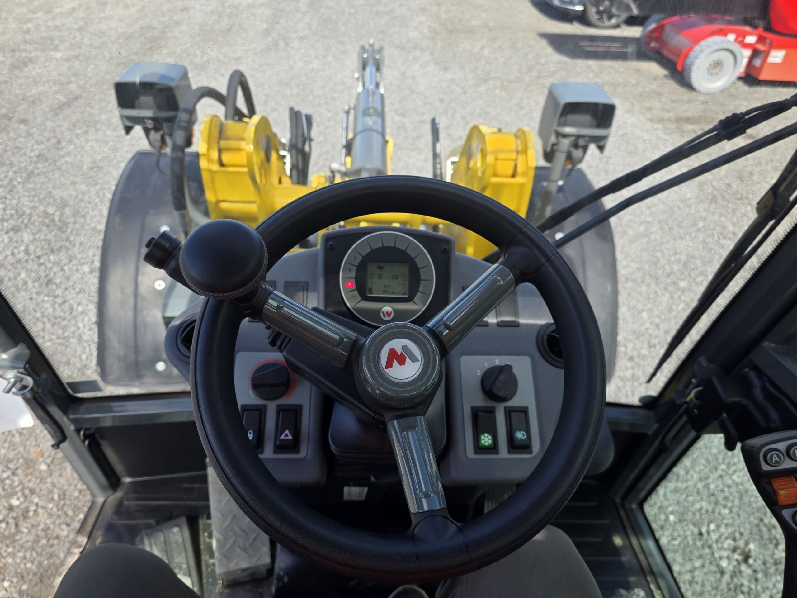 Vue depuis le siège conducteur d'un engin de chantier montrant le volant, les boutons de commande et le tableau de bord doté d'un écran numérique. Les bras jaunes de l'engin sont visibles à travers le pare-brise.