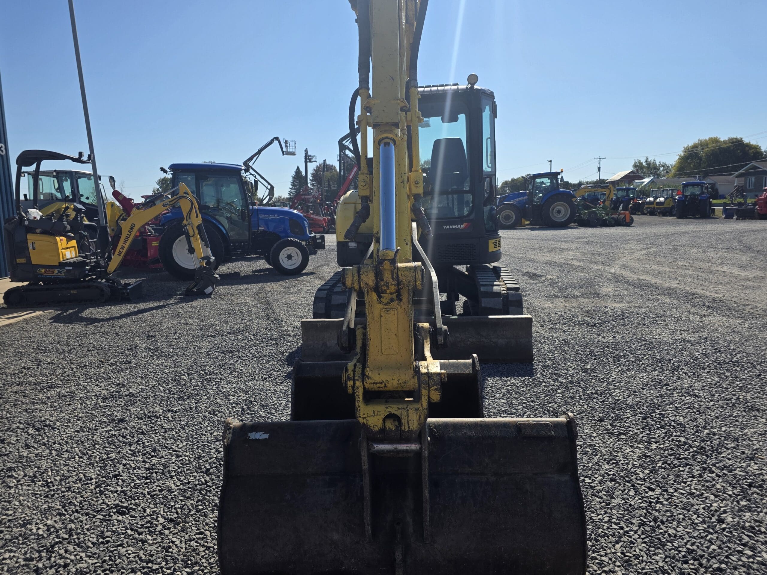 Une pelleteuse jaune est garée sur un terrain en gravier, face à la caméra ; on aperçoit à l'arrière-plan d'autres engins de chantier et des tracteurs, par une journée ensoleillée.