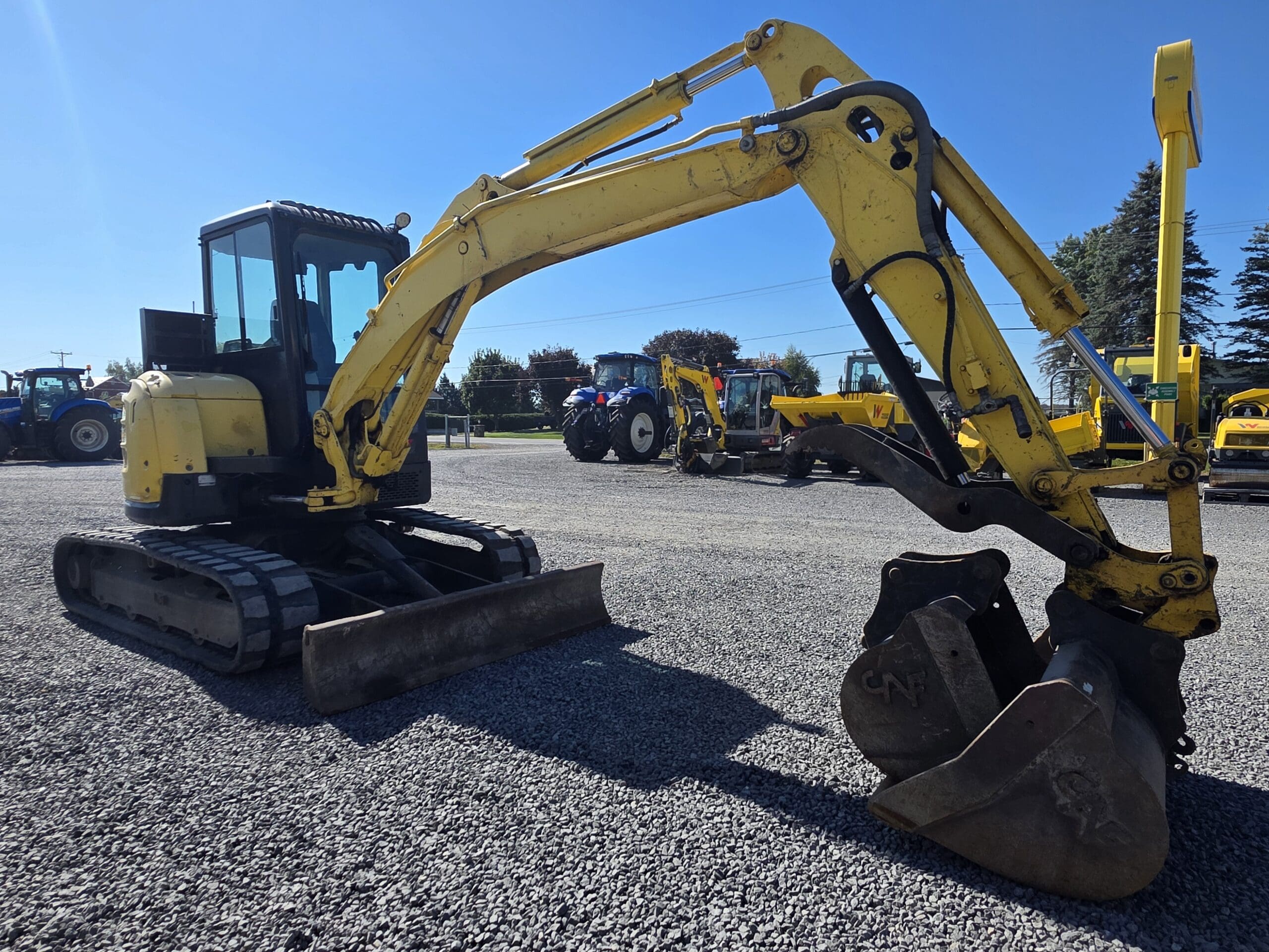 Une pelleteuse jaune équipée de deux godets est garée sur un terrain en gravier, entourée d’autres engins de chantier, sous un ciel bleu limpide.