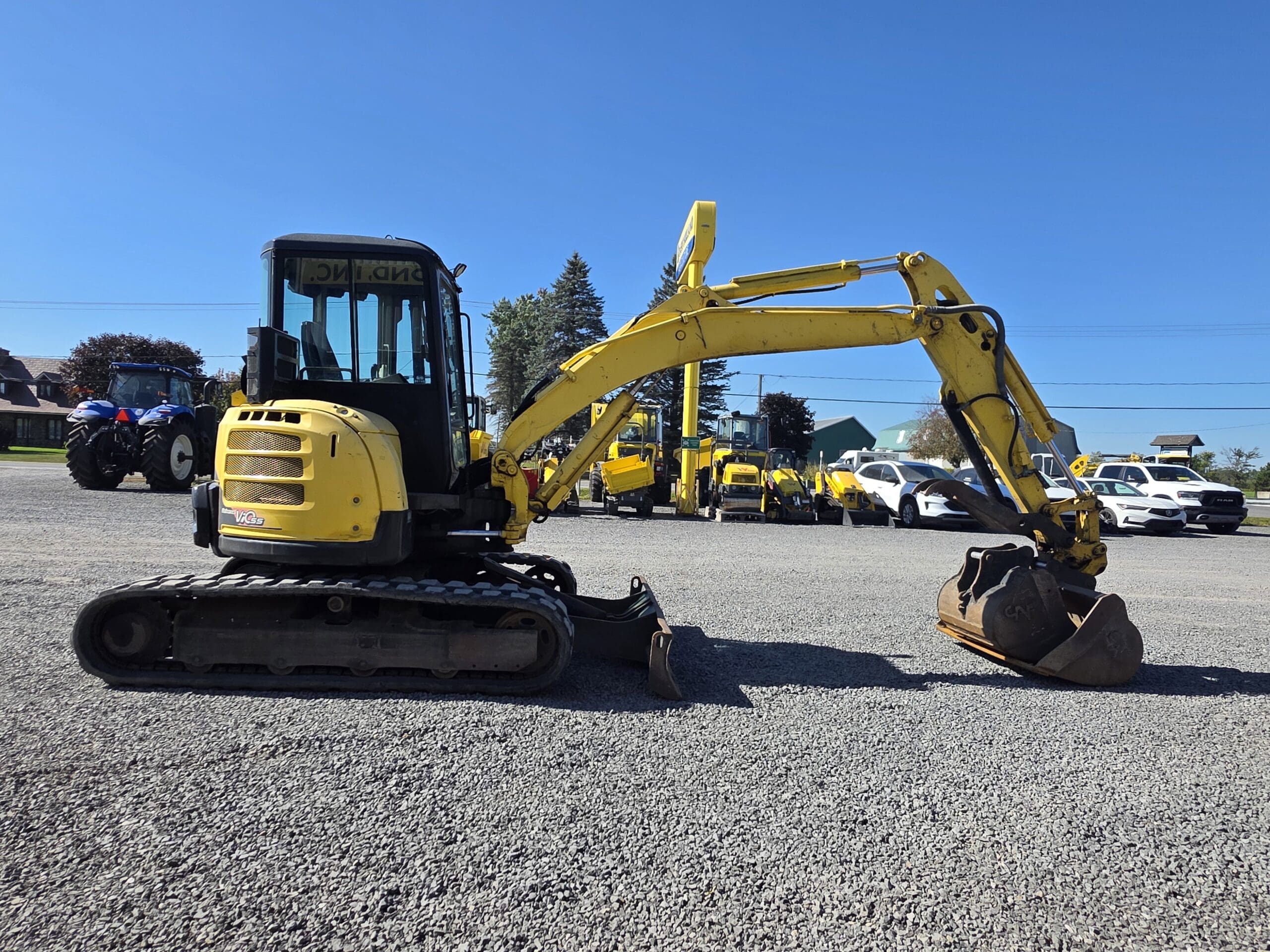 Une mini-pelle jaune équipée d'un godet est garée sur du gravier. D'autres engins de chantier et des voitures blanches sont visibles à l'arrière-plan, sous un ciel bleu limpide.