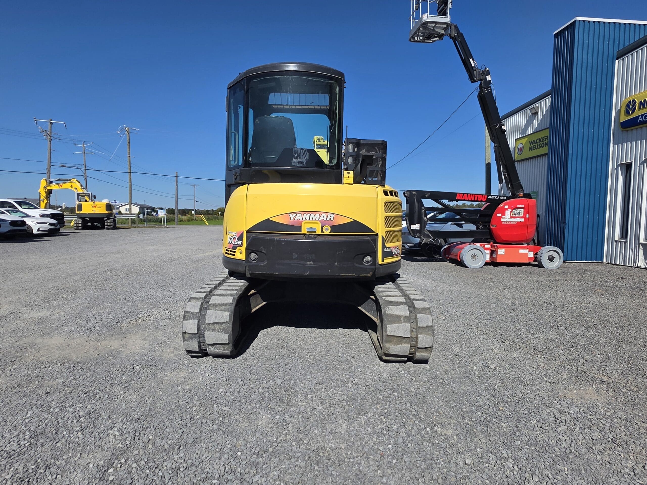 Une pelleteuse Yanmar jaune est garée sur du gravier près d'un bâtiment bleu. D'autres engins de chantier, des voitures et des poteaux électriques sont visibles à l'arrière-plan, sous un ciel bleu limpide.